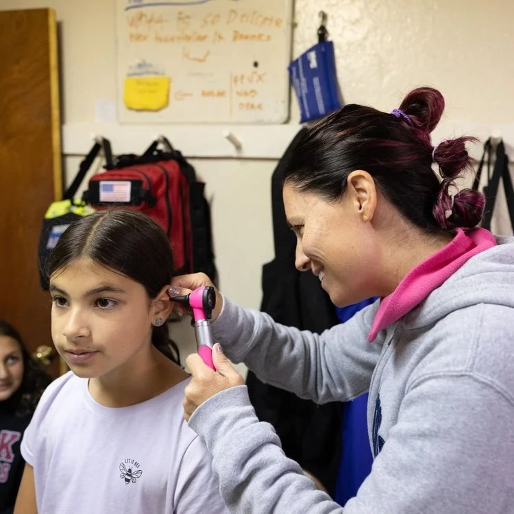 Woman using an electronic ear cleaning device on a girl in a classroom.