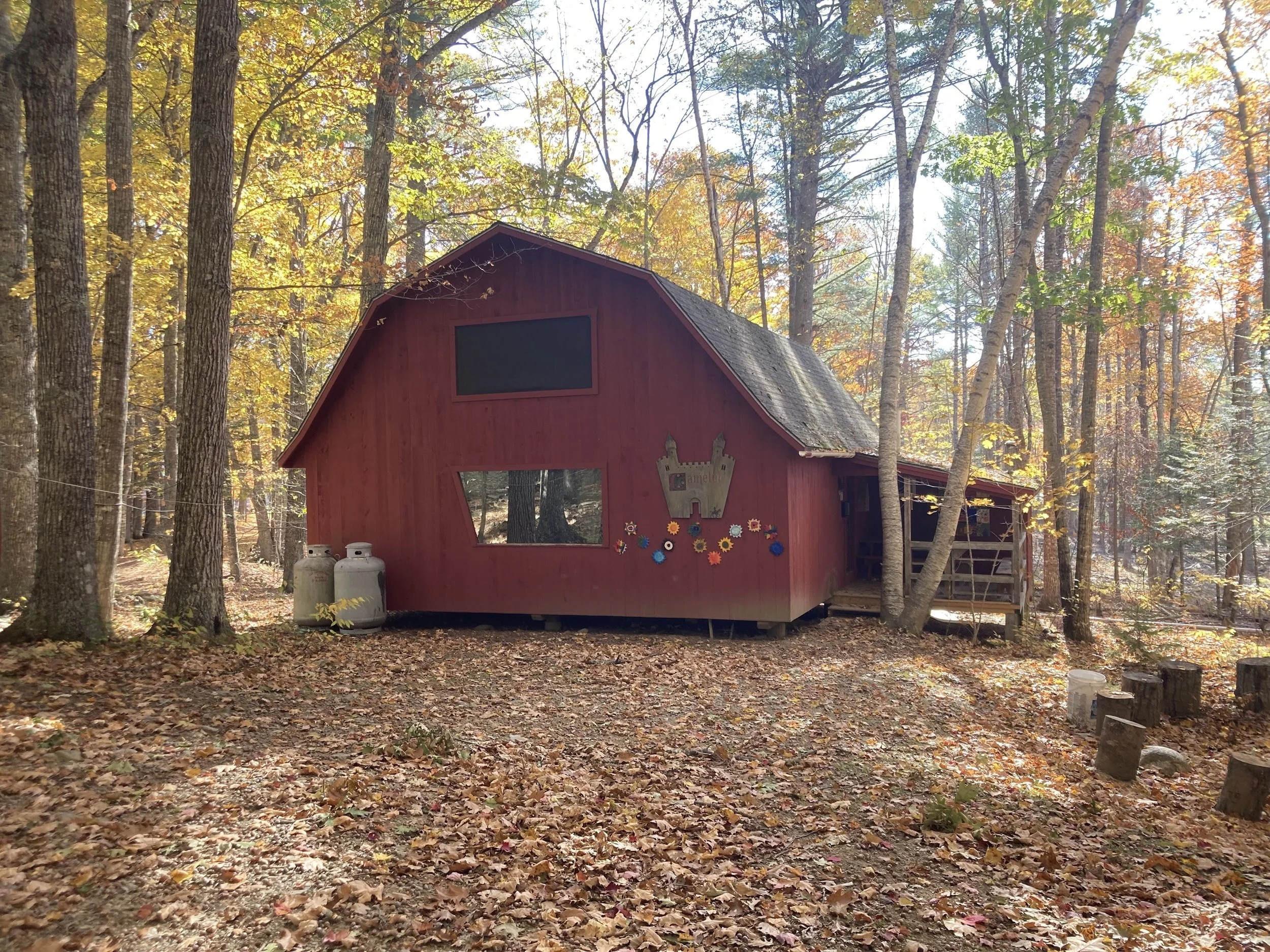 A red house-like structure in the woods with colorful decorations on the wall, surrounded by trees with fall foliage.