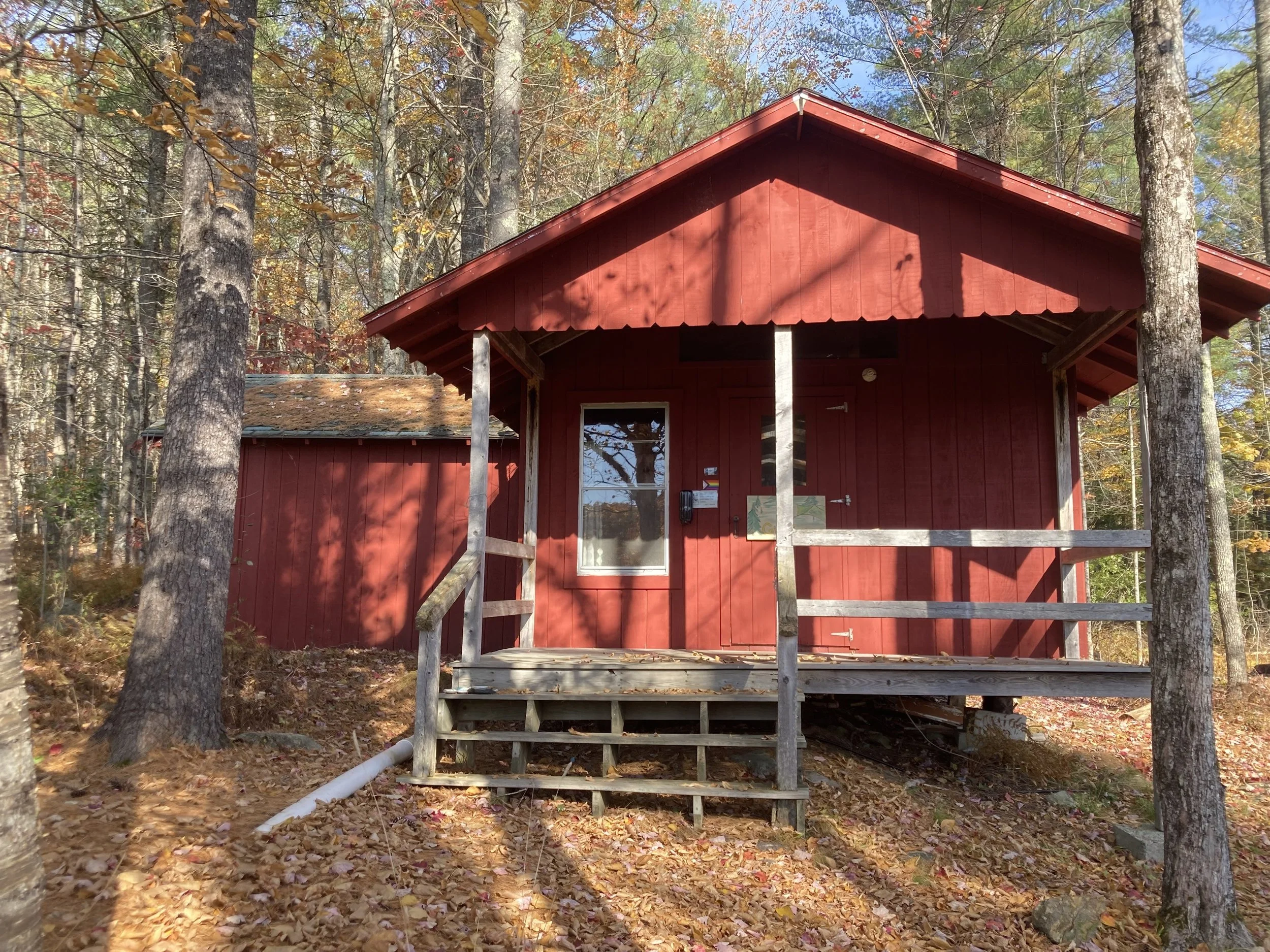 A small red wooden cabin with a porch, surrounded by trees with fall foliage.