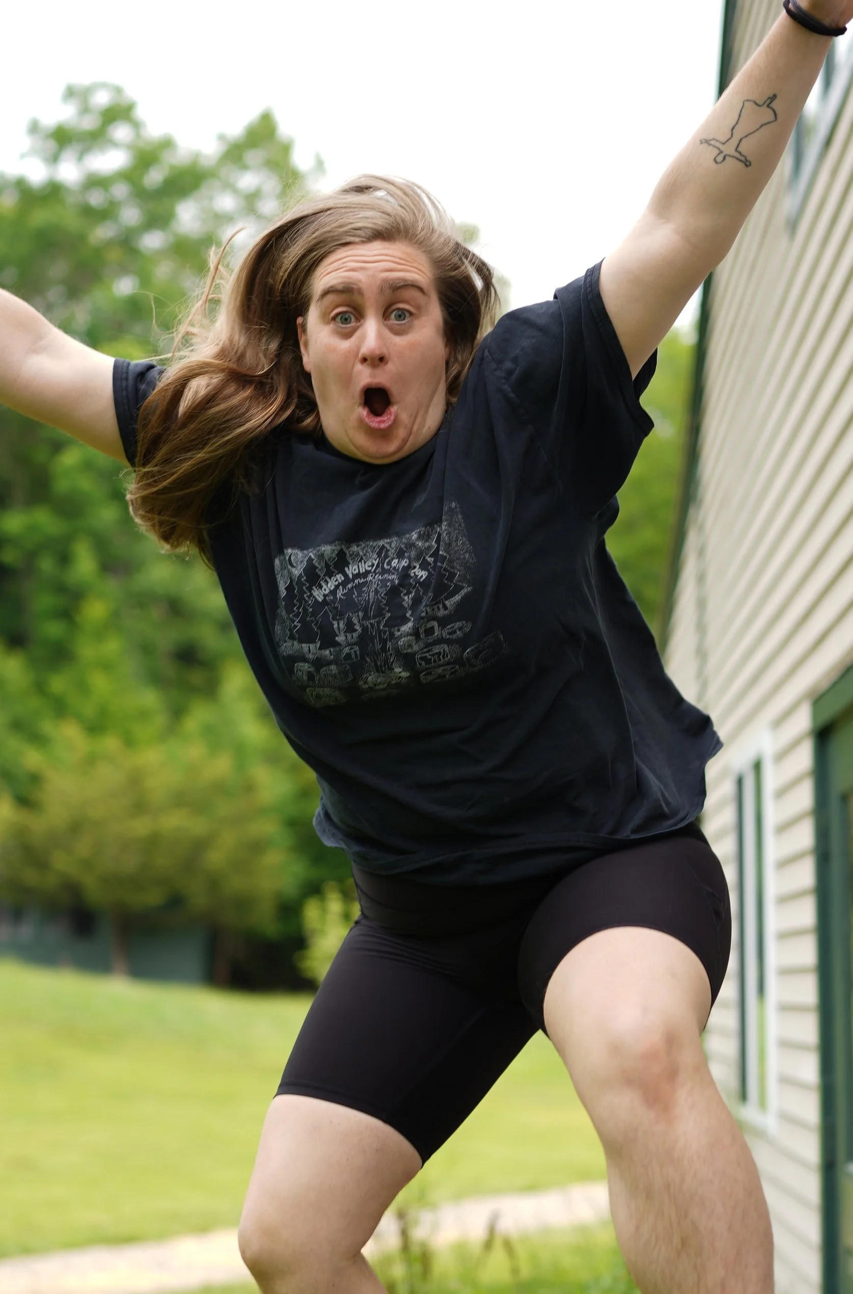 Person outdoors, wearing black shorts and a black t-shirt, mid-movement, with a surprised or intense expression, and a tattoo of a map outline on their right arm, on a background of green trees and a building.