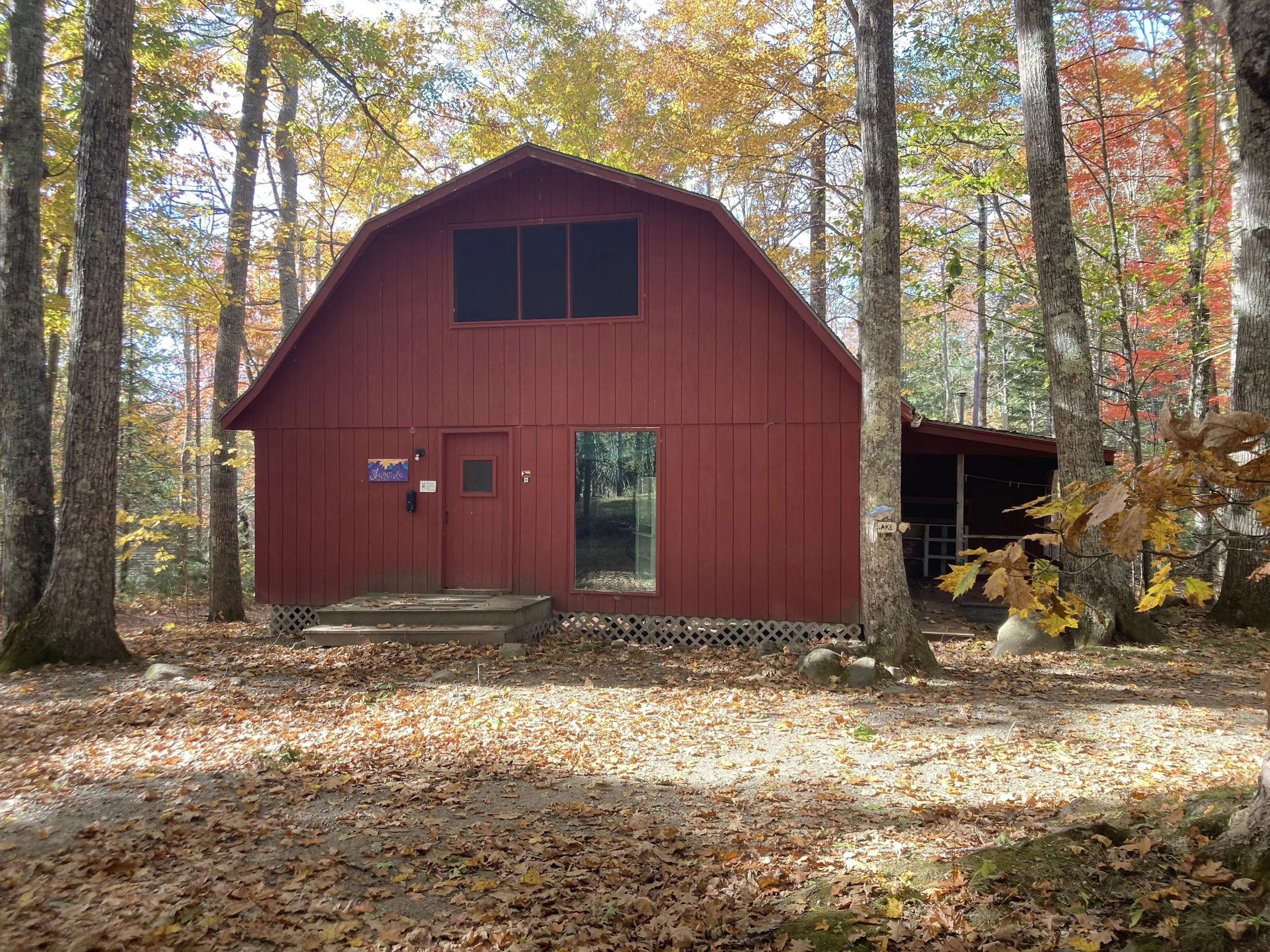Red cabin in a wooded forest during fall with colorful leaves on the ground and on the trees.