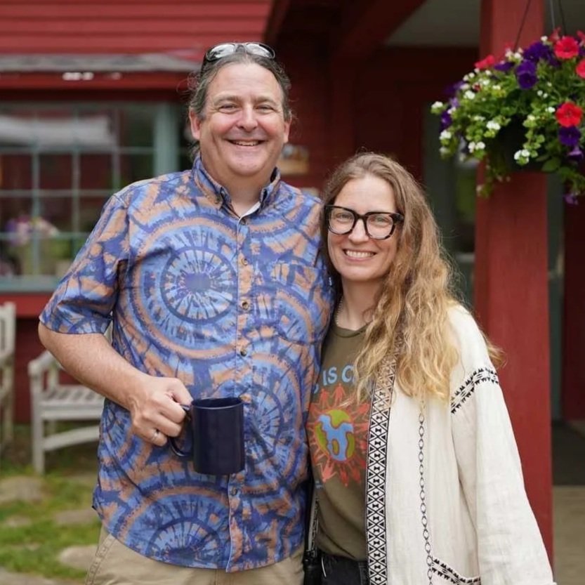 Two smiling people, a man and a woman, standing outdoors in front of a red building with a hanging flower basket. The man is holding a blue mug, and the woman is wearing glasses and a colorful t-shirt with a white cardigan.