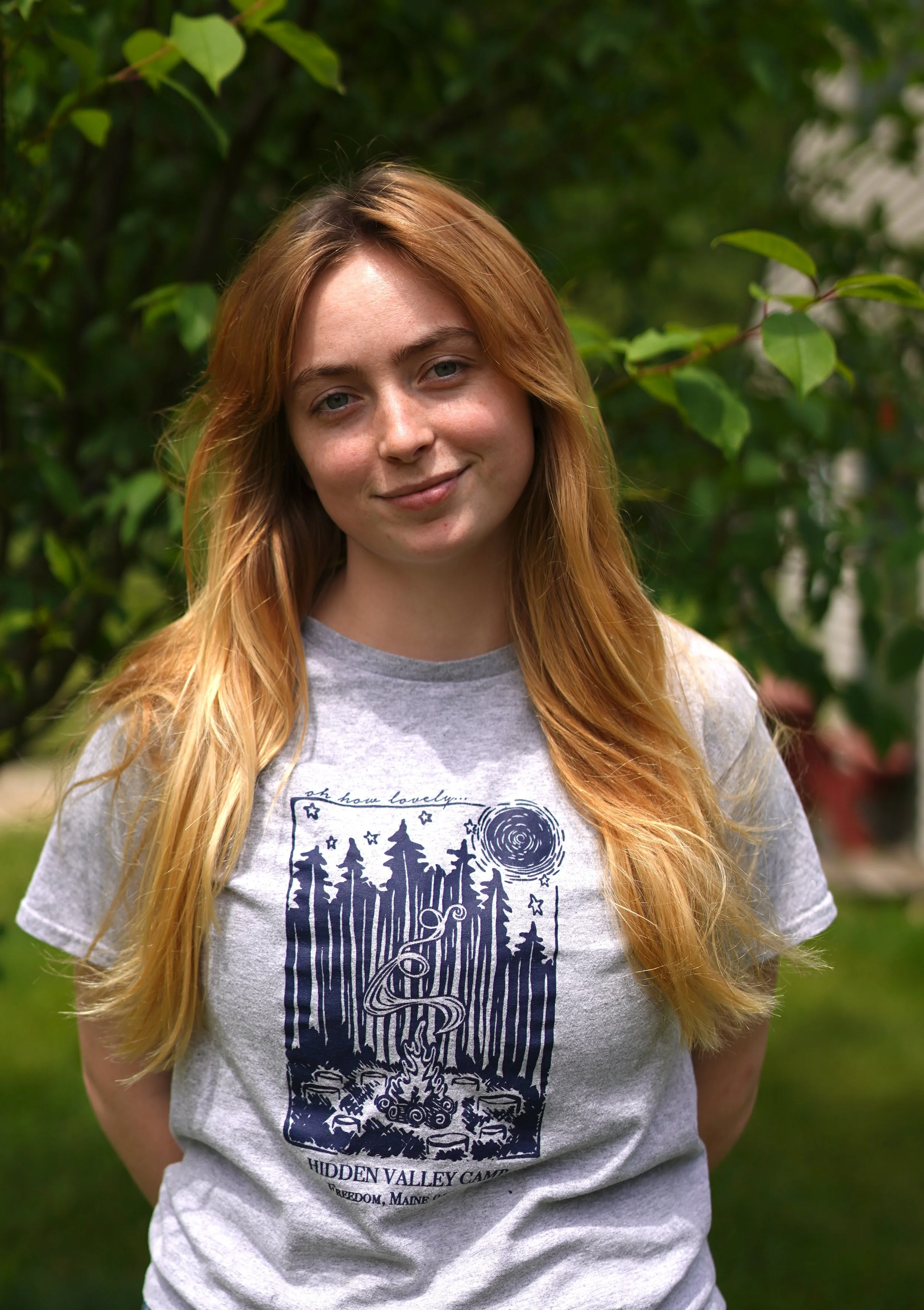 A young woman with long, light brown hair and fair skin standing outdoors in front of green foliage. She is wearing a gray T-shirt with a graphic design and text that reads "Hidden Valley Camp, Freedom, Maine." She is smiling gently.