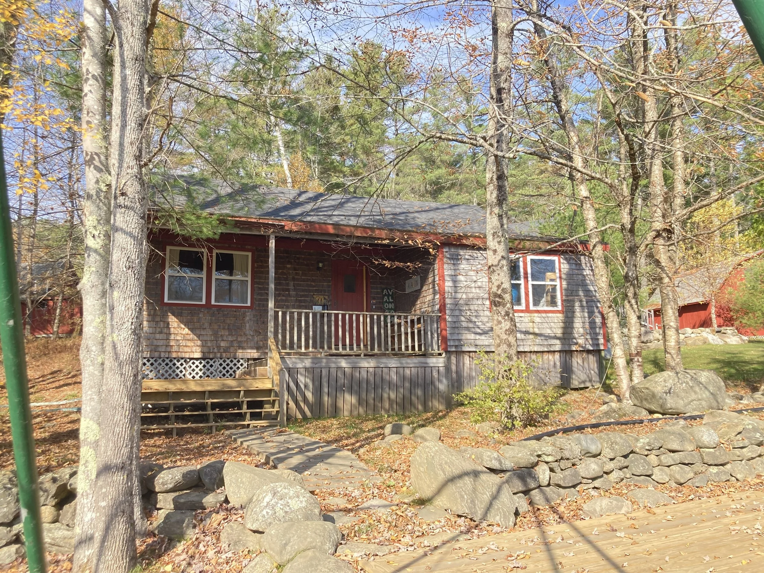 A small wooden house with a front porch and steps, surrounded by trees with fall foliage, rocks, and a stone wall in front.