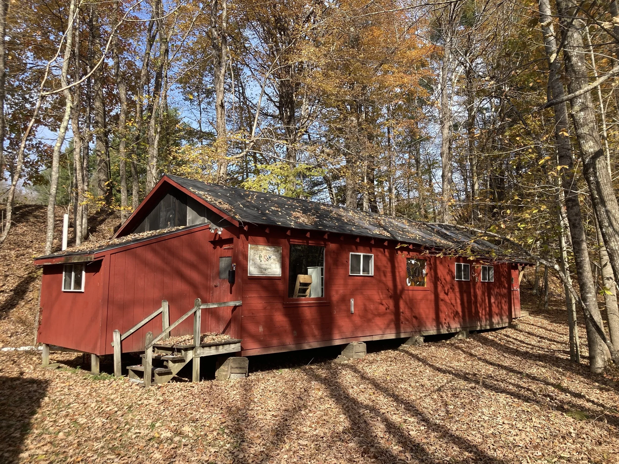 A red wooden cabin with a sloped roof, windows, and a small staircase leading to the door, situated in a wooded area with autumn leaves on the ground and trees with yellow, orange, and green foliage.