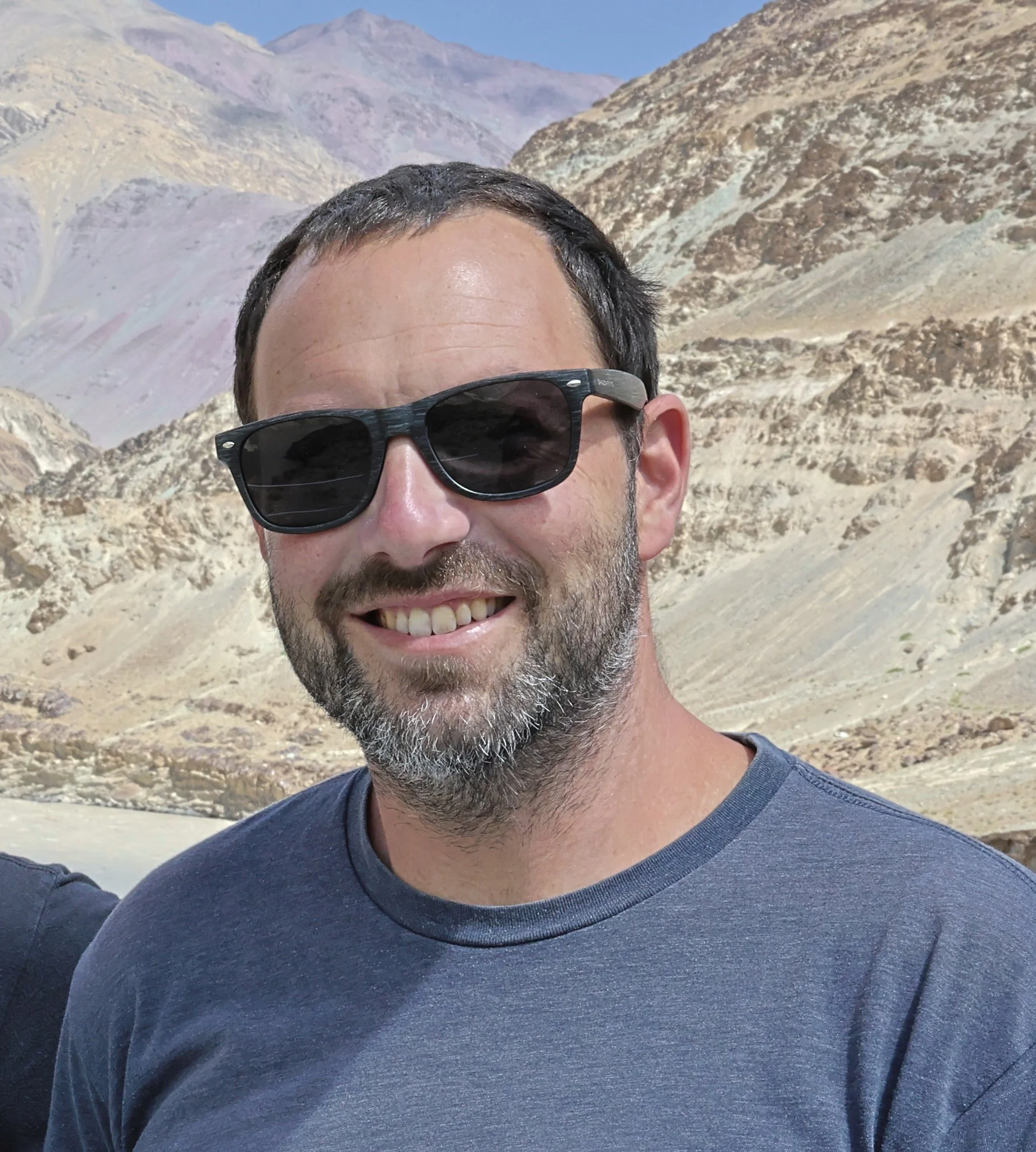 Close-up photo of a smiling man with dark hair, wearing black sunglasses and a dark blue T-shirt, in a desert landscape with mountains in the background.