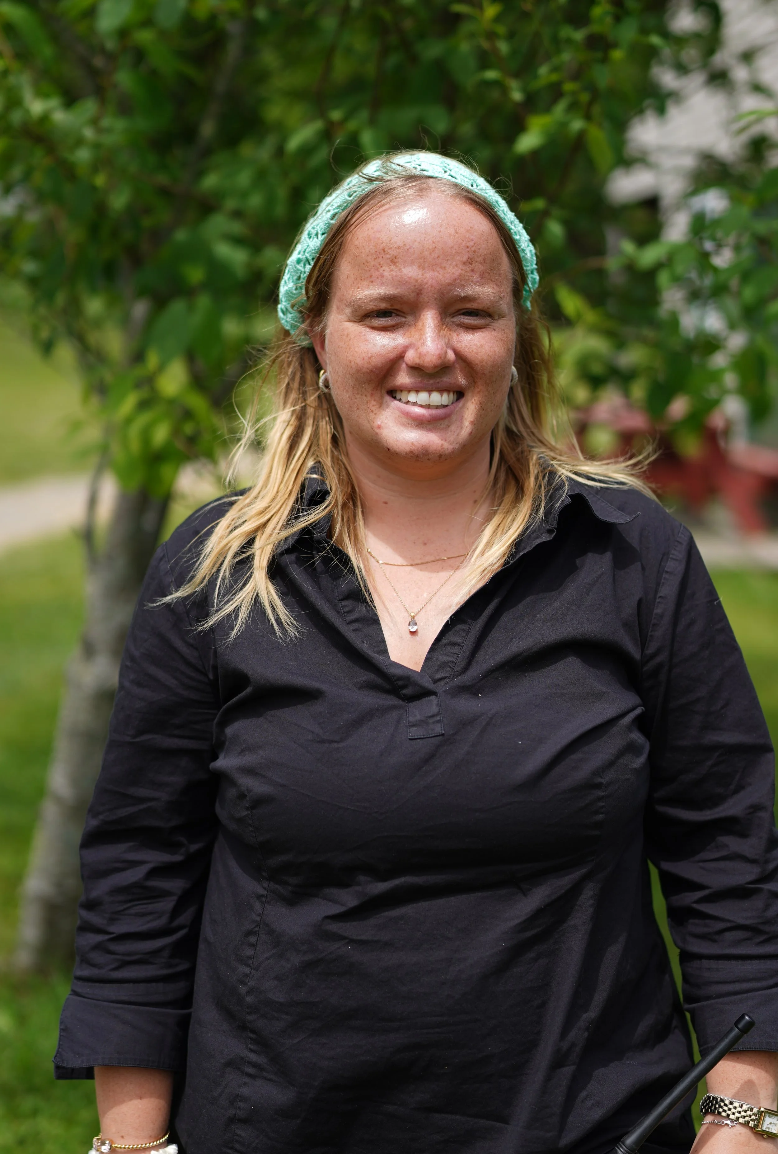 A woman with blonde hair wearing a pastel green headband and a black shirt, smiling outdoors with greenery in the background.