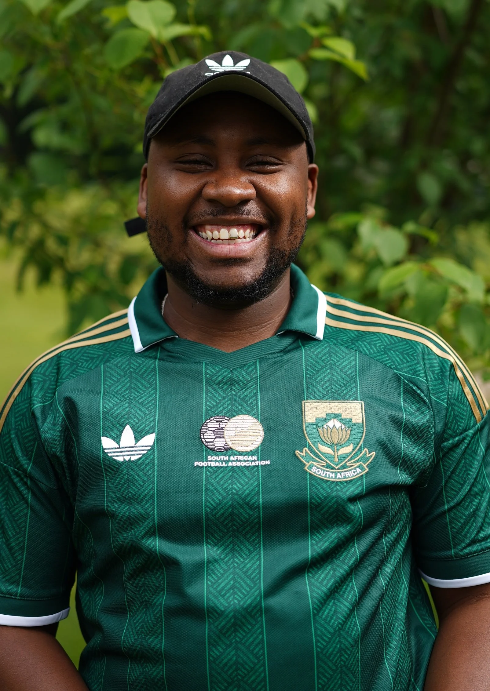 Close-up of a smiling man wearing a South African football jersey and cap, standing outdoors with green foliage in the background.