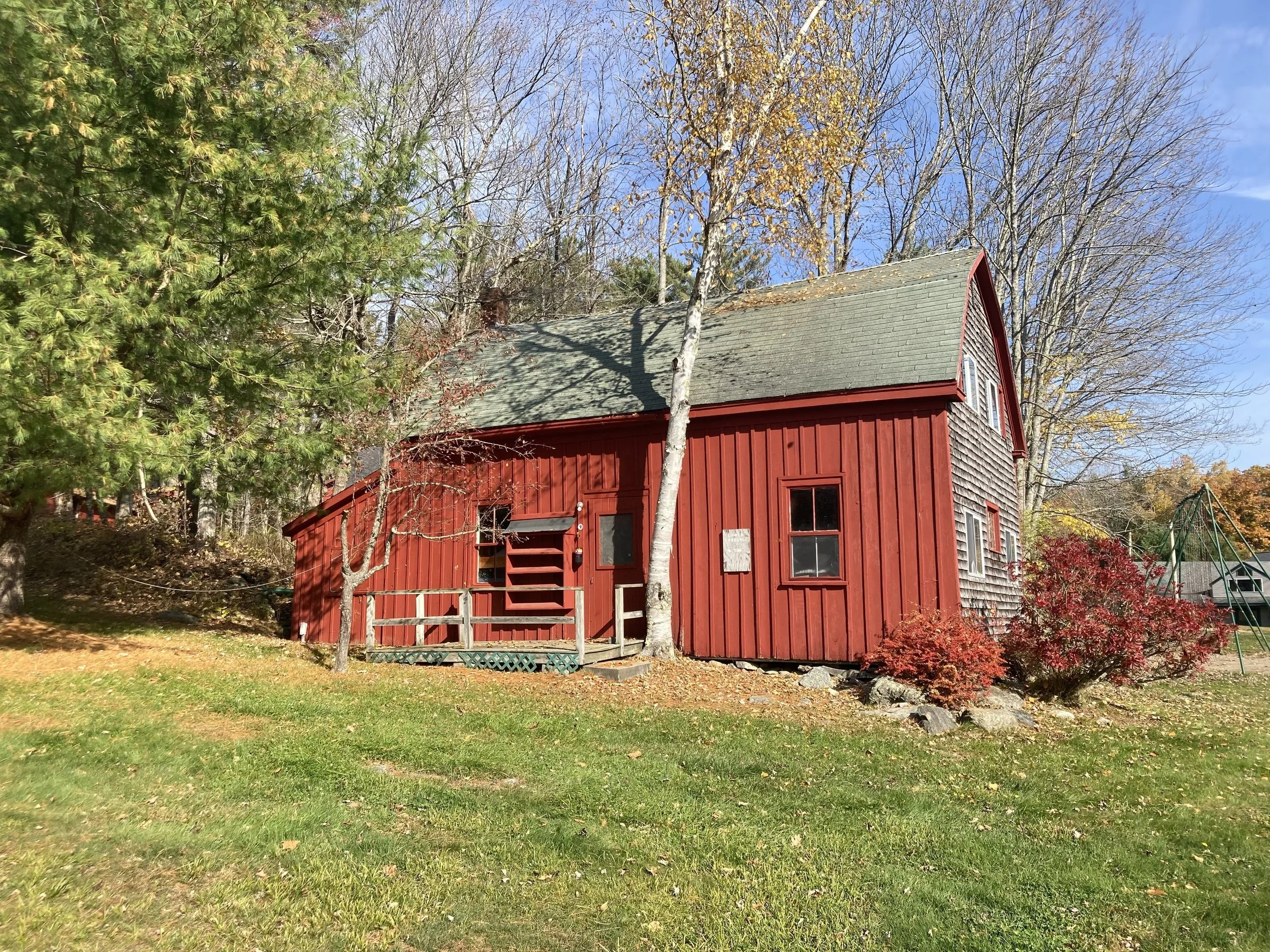 A red wooden barn with a gray shingled roof, surrounded by trees and a grassy yard, with some autumn foliage and a clear blue sky.