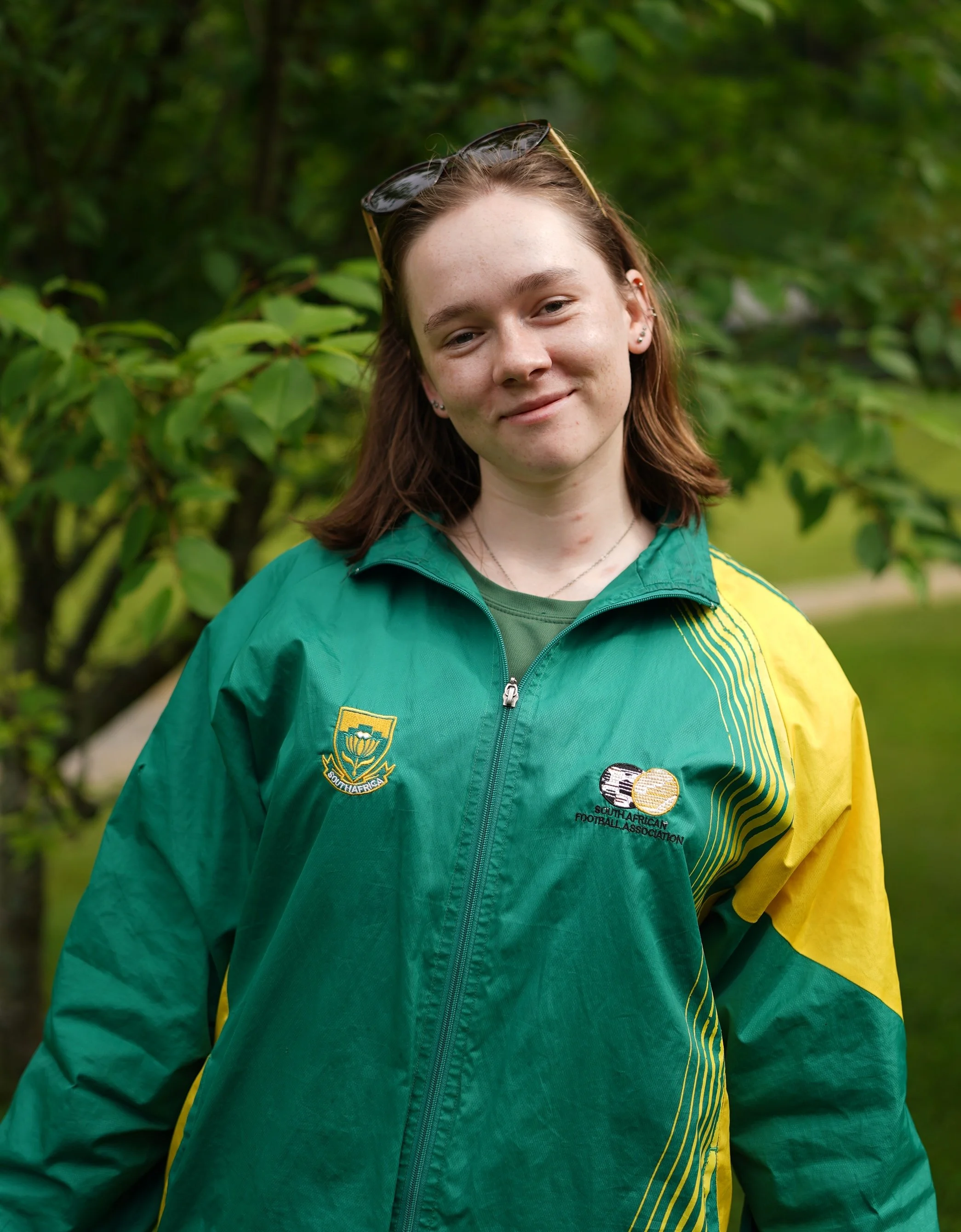 A young woman standing outdoors in front of green foliage, wearing a green and yellow South African Football Association jacket and sunglasses on her head.