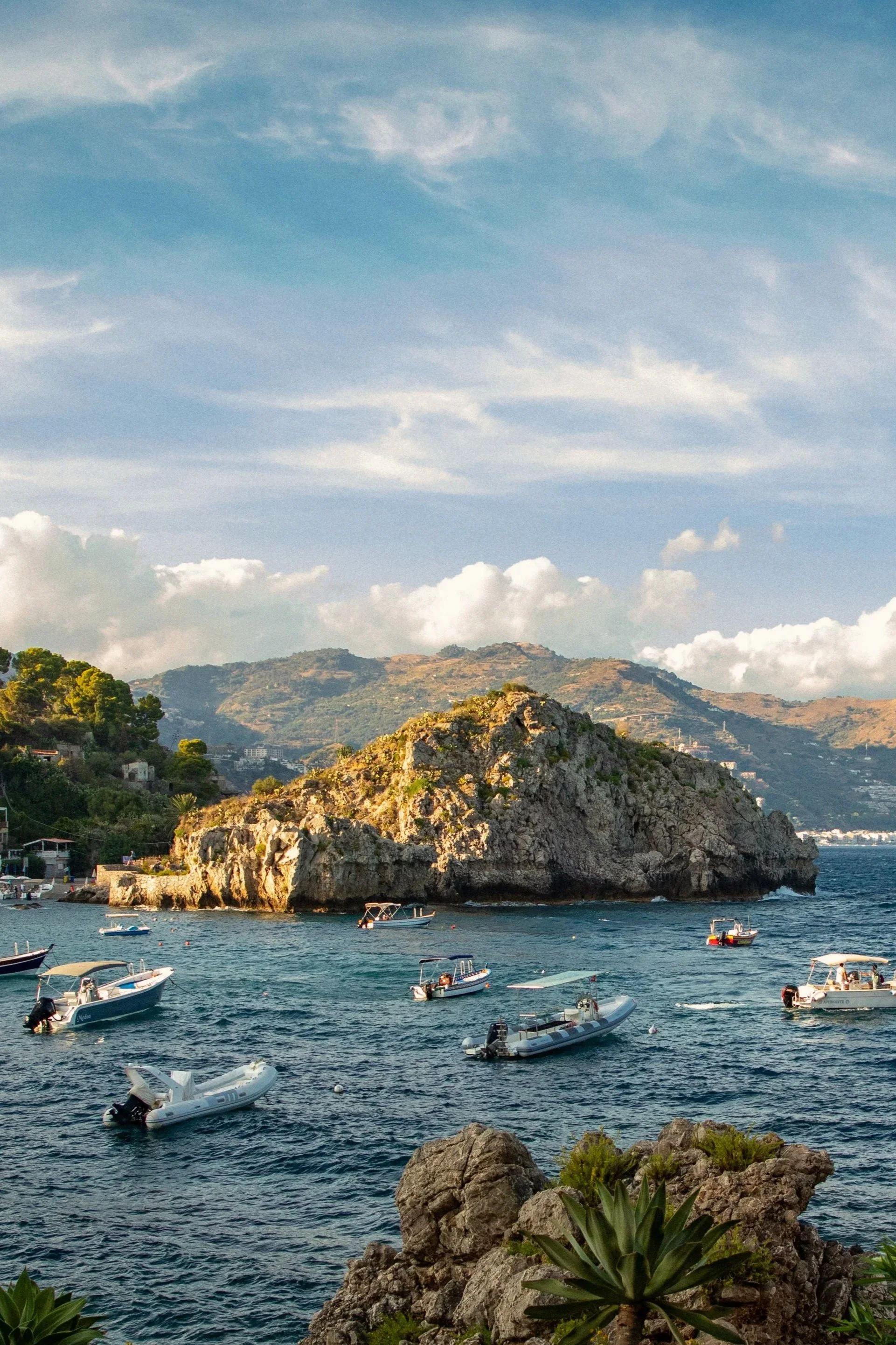 A coastal scene with boats on the water, rocky shoreline, a large rock formation, green hills in the background, and partly cloudy sky. Sicily views, Italian best travel. Hotel Belmond Sant Andrea
