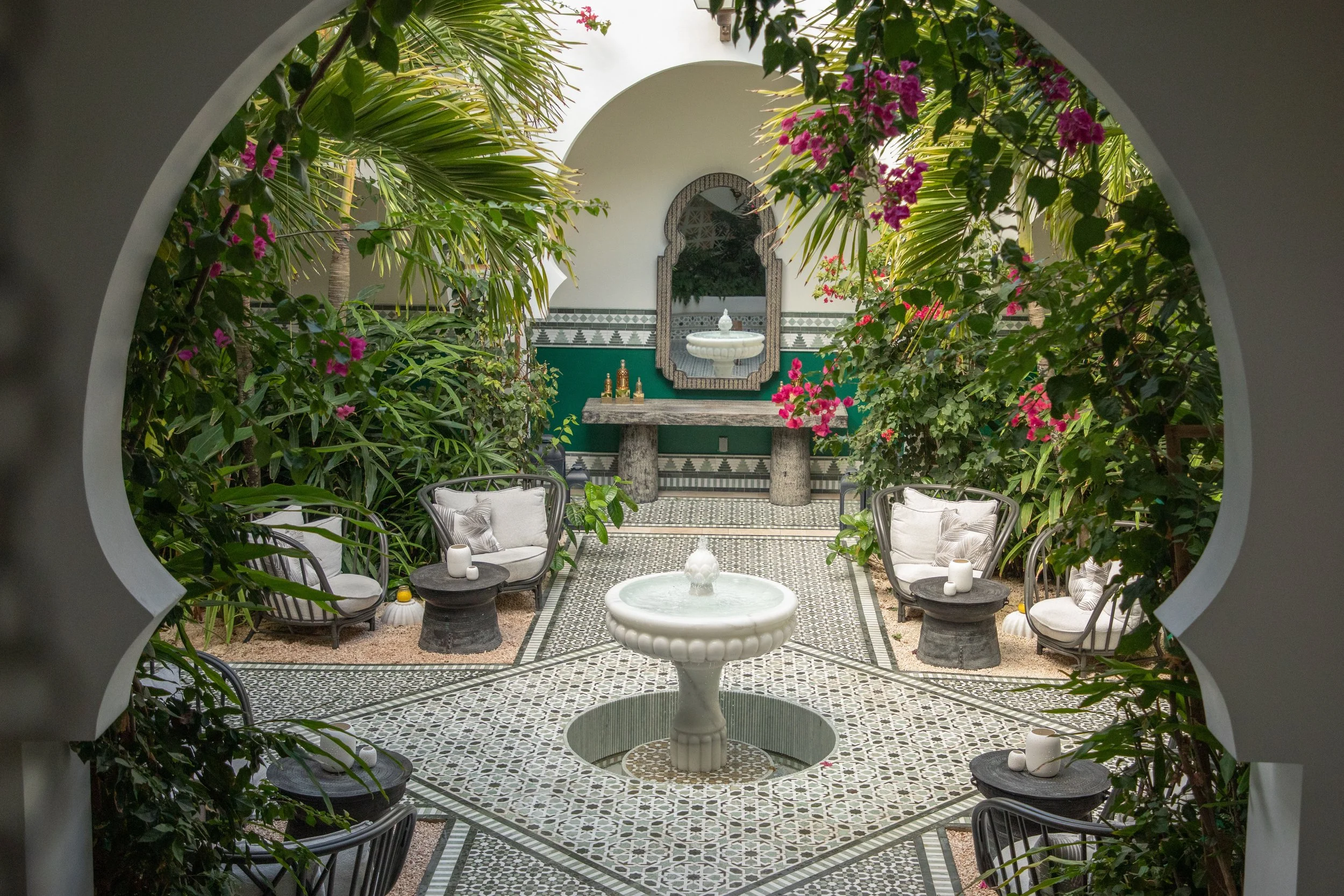 Indoor courtyard with patterned tile floor, greenery, pink flowers, white seating with pillows, small black tables with candles, and a fountain in the center, framed by an arched opening. Guerlain luxury spa at the caribbean hotel Belmond Cap Juluca.