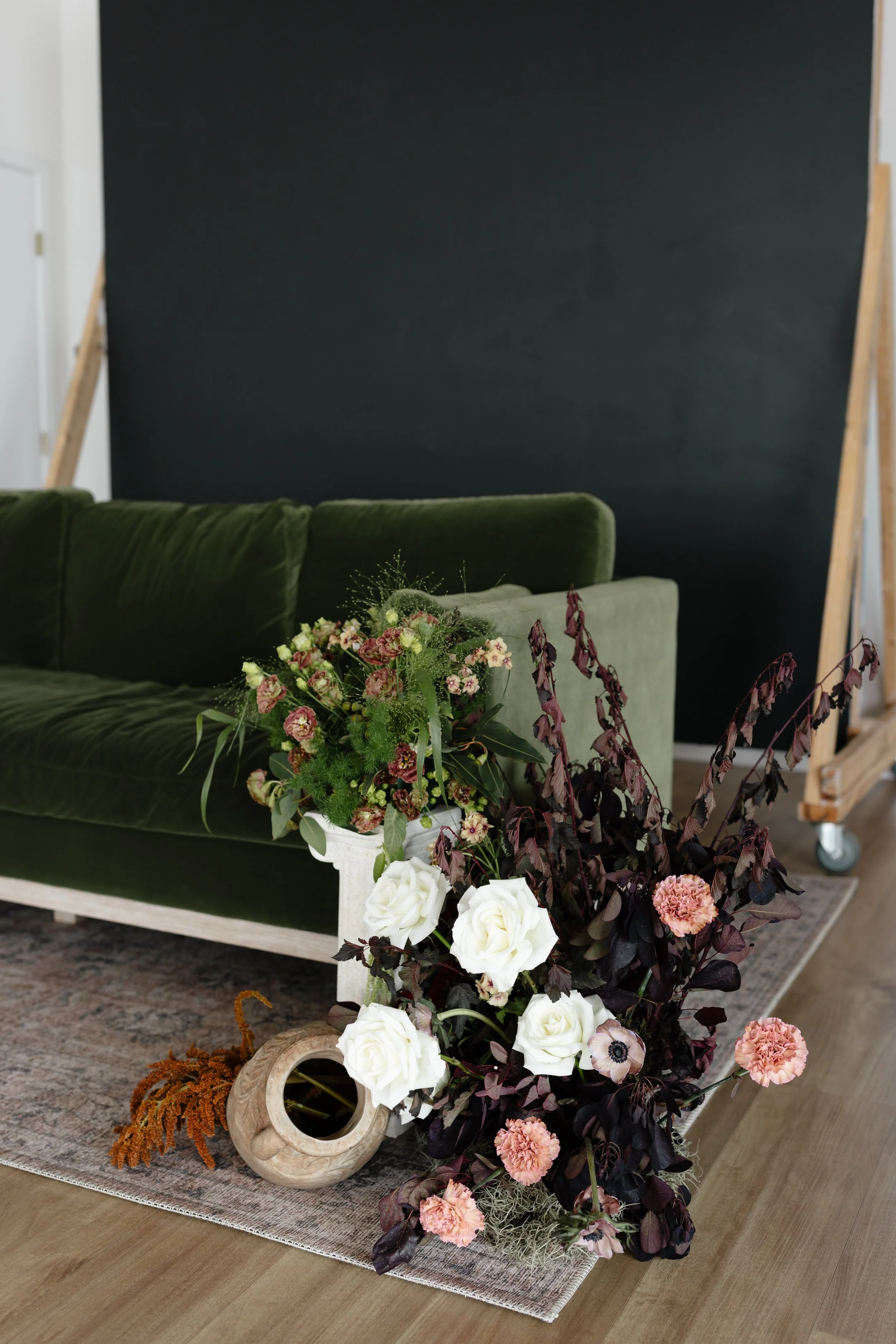A floral arrangement with white roses, pink carnations, and green and dark purple foliage, placed on a beige rug in front of a green velvet sofa and a dark chalkboard wall.
