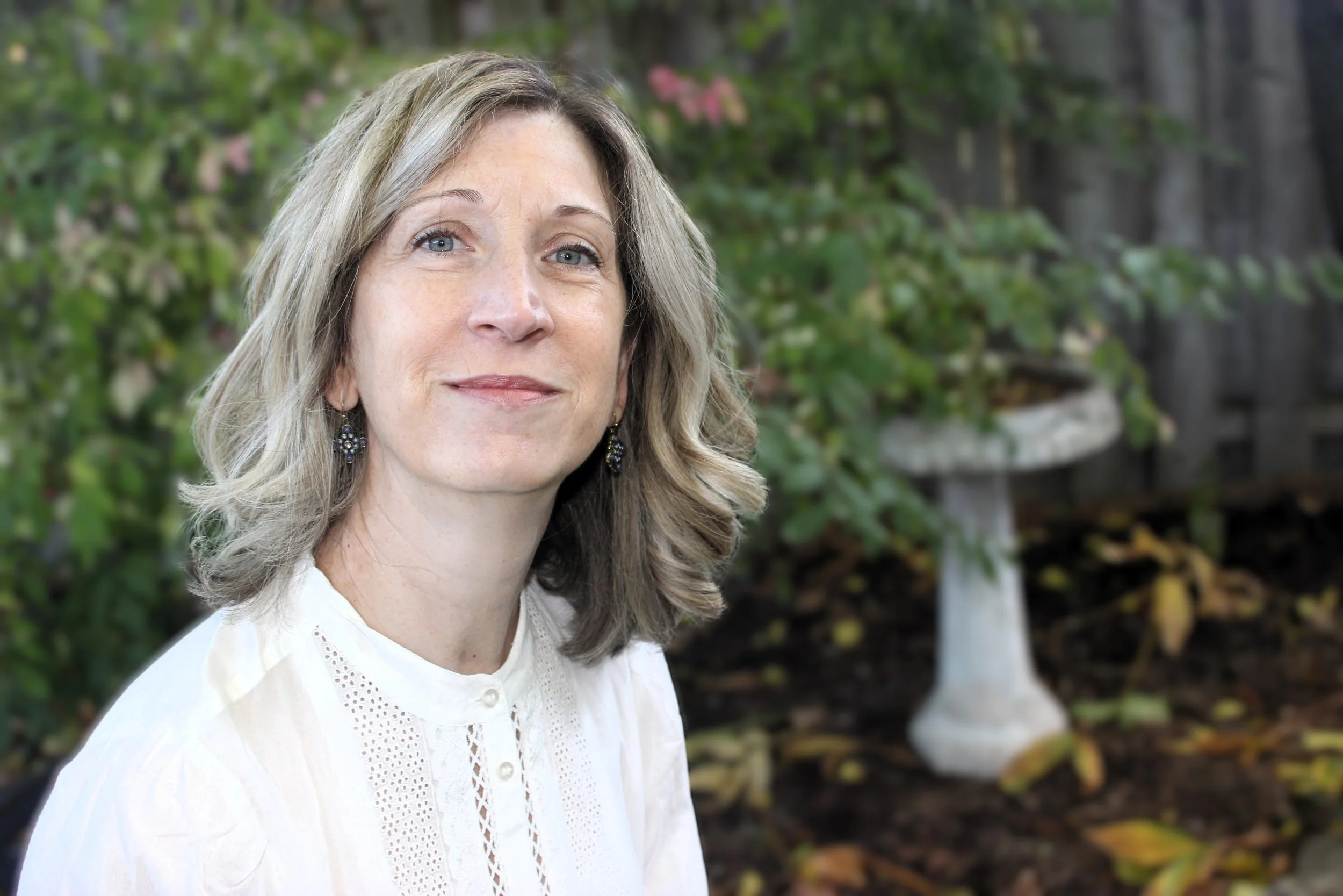 A middle-aged woman with shoulder-length light brown hair, wearing a white blouse with decorative embroidery, posing outdoors with a blurred garden background.