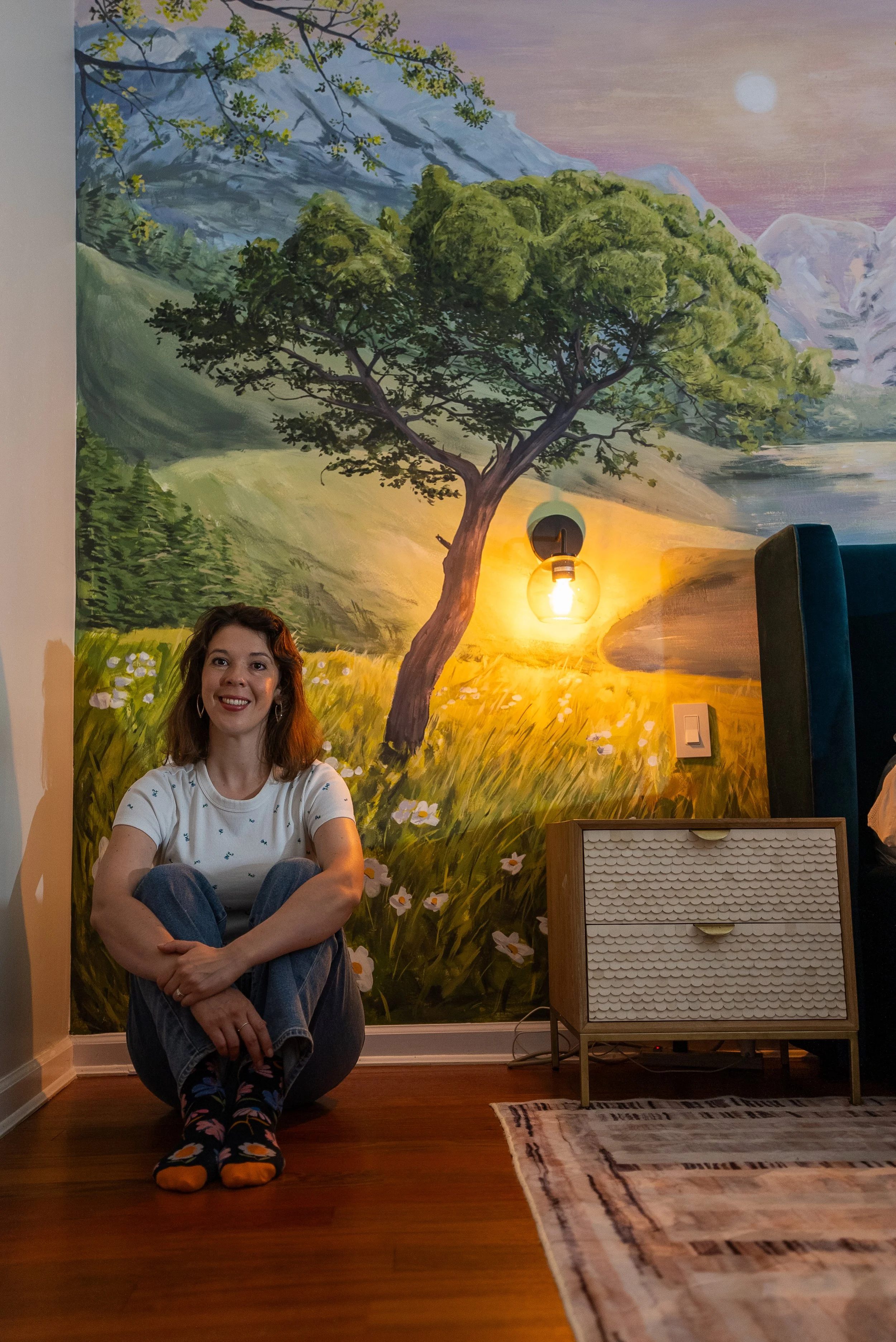 A woman with red hair sitting on a wooden floor in front of a large landscape mural. The mural depicts a mountain scene with a large green tree, a sunset or sunrise, and snow-capped peaks in the background. There is a wall with a light switch, a wooden dresser, a patterned rug, and a wall-mounted light bulb with a warm glow.