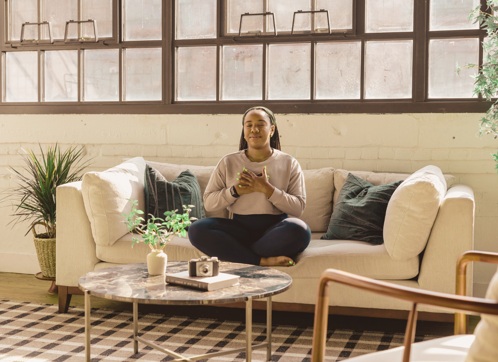 BIPOC Woman sitting with her eyes closed and hands over her heart in a calm, sunlit space.