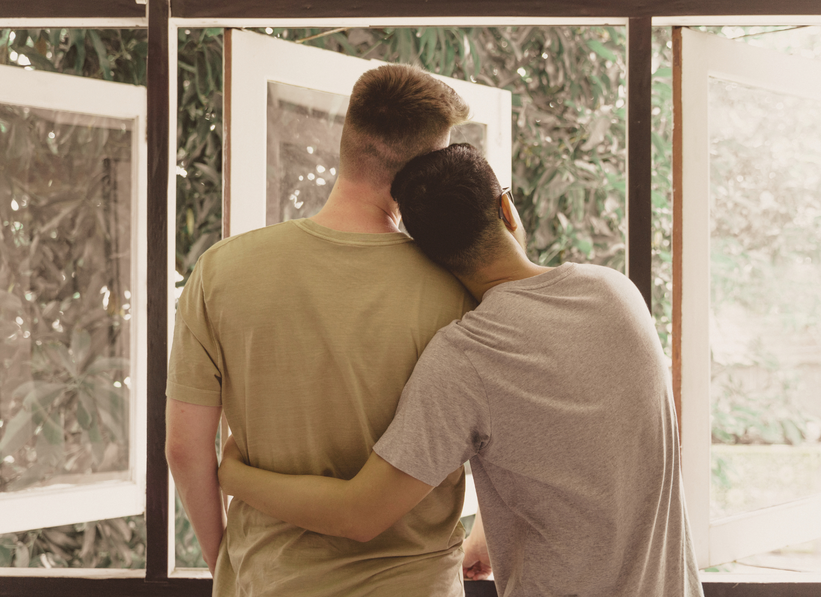 Two men standing together by a window, one resting his head on the other's shoulder