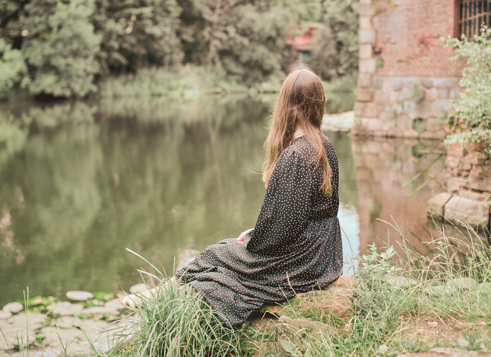 Woman sitting beside a peaceful pond surrounded by greenery.