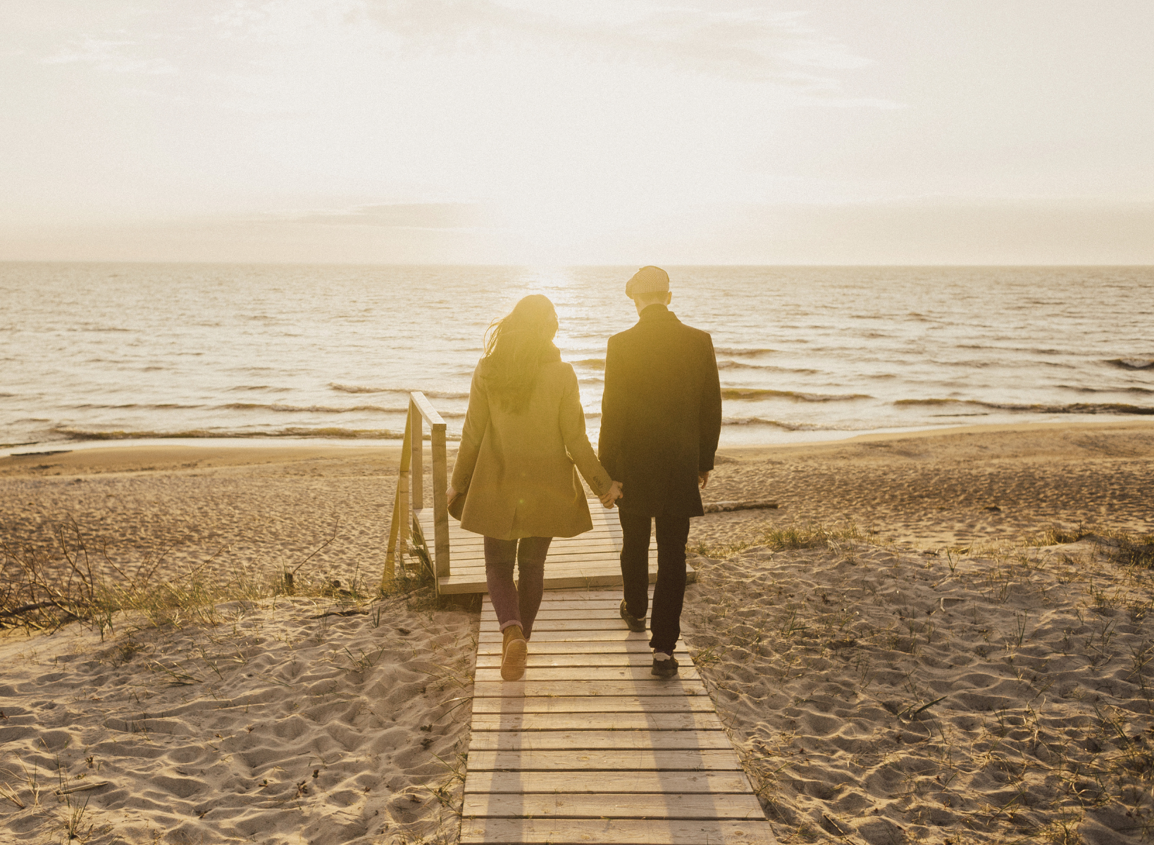 Couple holding hands as they walk toward the beach along a wooden boardwalk