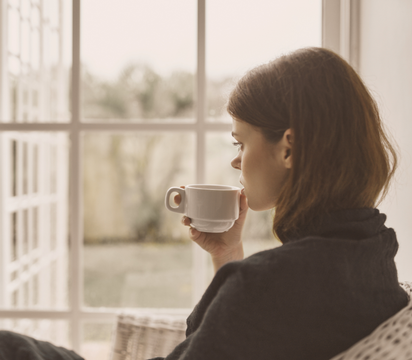 Person holding a cup while sitting quietly beside a large window.