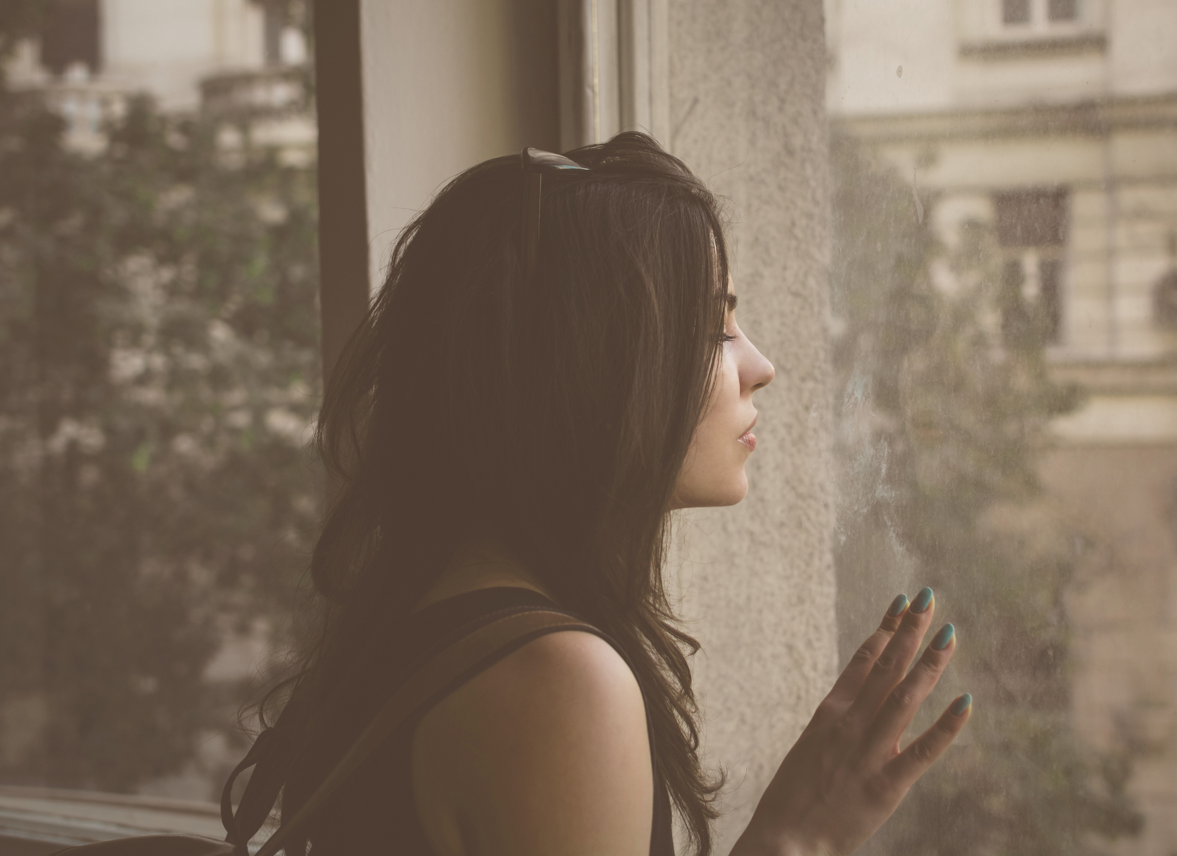 Person standing by a window, looking outside with a hand resting against the glass.