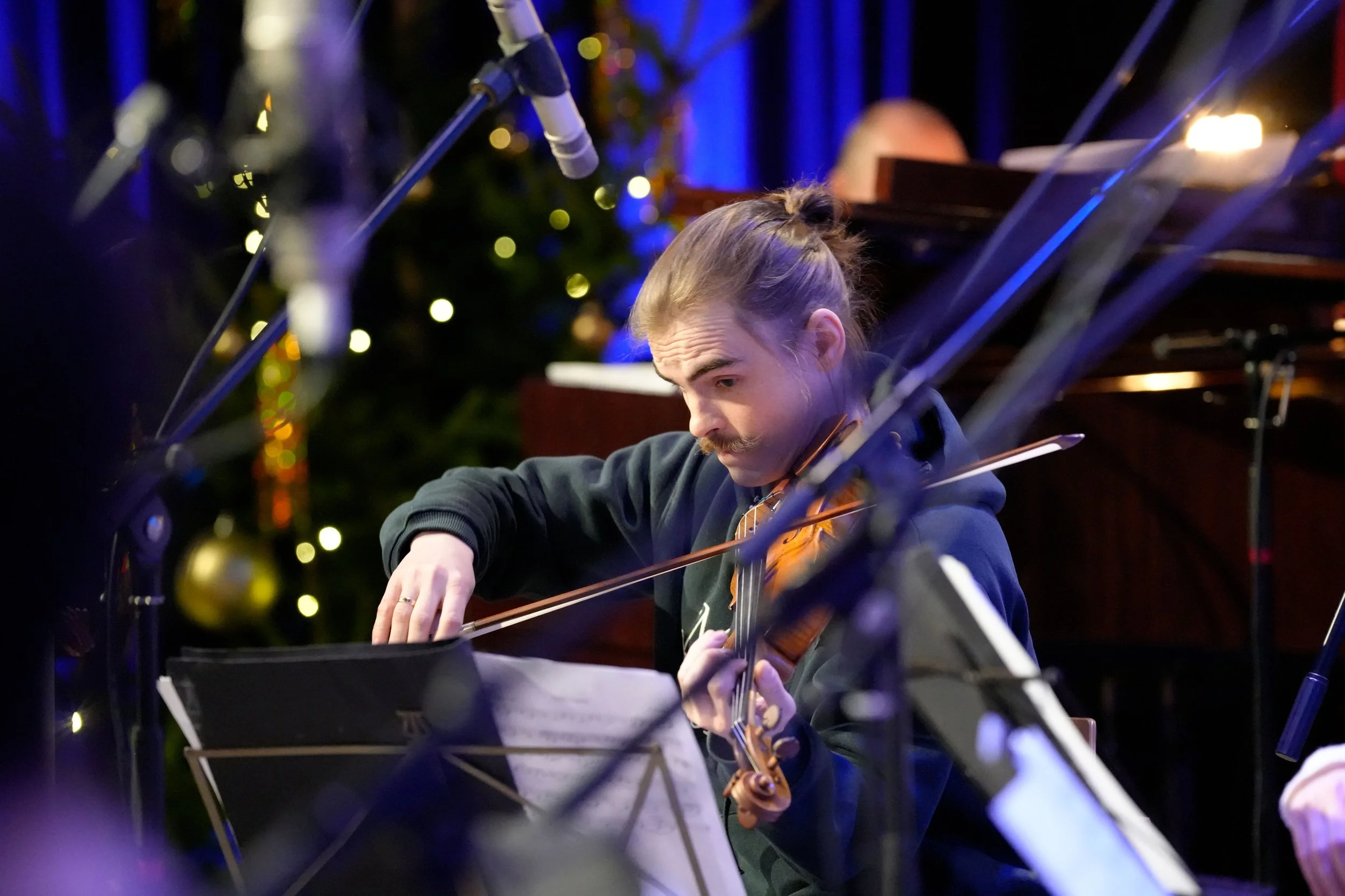 Mann spielt Geige bei einem Konzert, umgeben von Musikständern und Mikrofonen, mit Weihnachtsdekoration im Hintergrund.