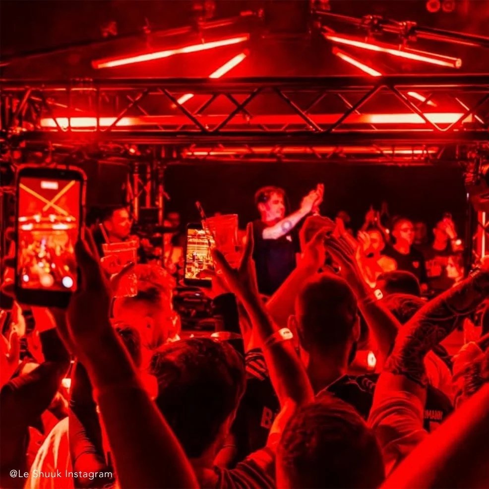 Menschen feiern auf einem Konzert mit roter Beleuchtung, einige nehmen Fotos auf, während ein DJ oder Performer auf der Bühne steht.