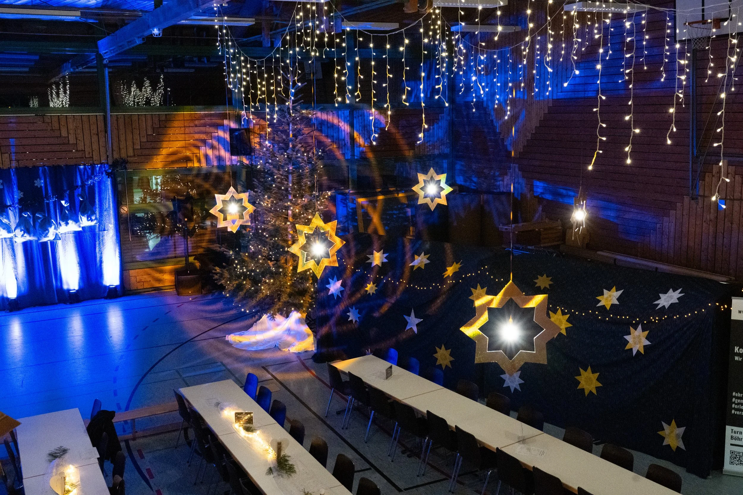 Innenraum mit Weihnachtsdekoration, Weihnachtsbaum, Lichterketten und festlicher Beleuchtung, Tische und Stühle.