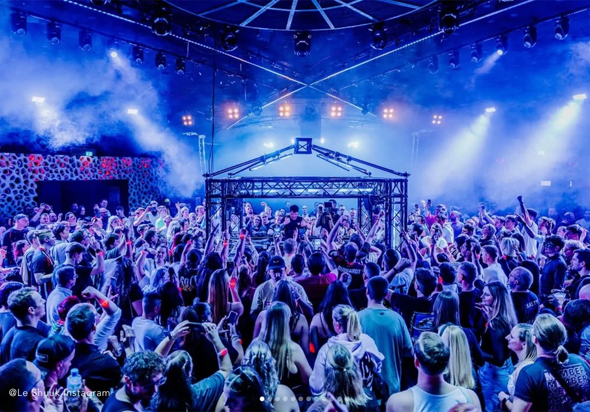 Menschenmenge bei einer elektronischen Musikveranstaltung mit DJ auf der Bühne, bunte Lichter und Nebel