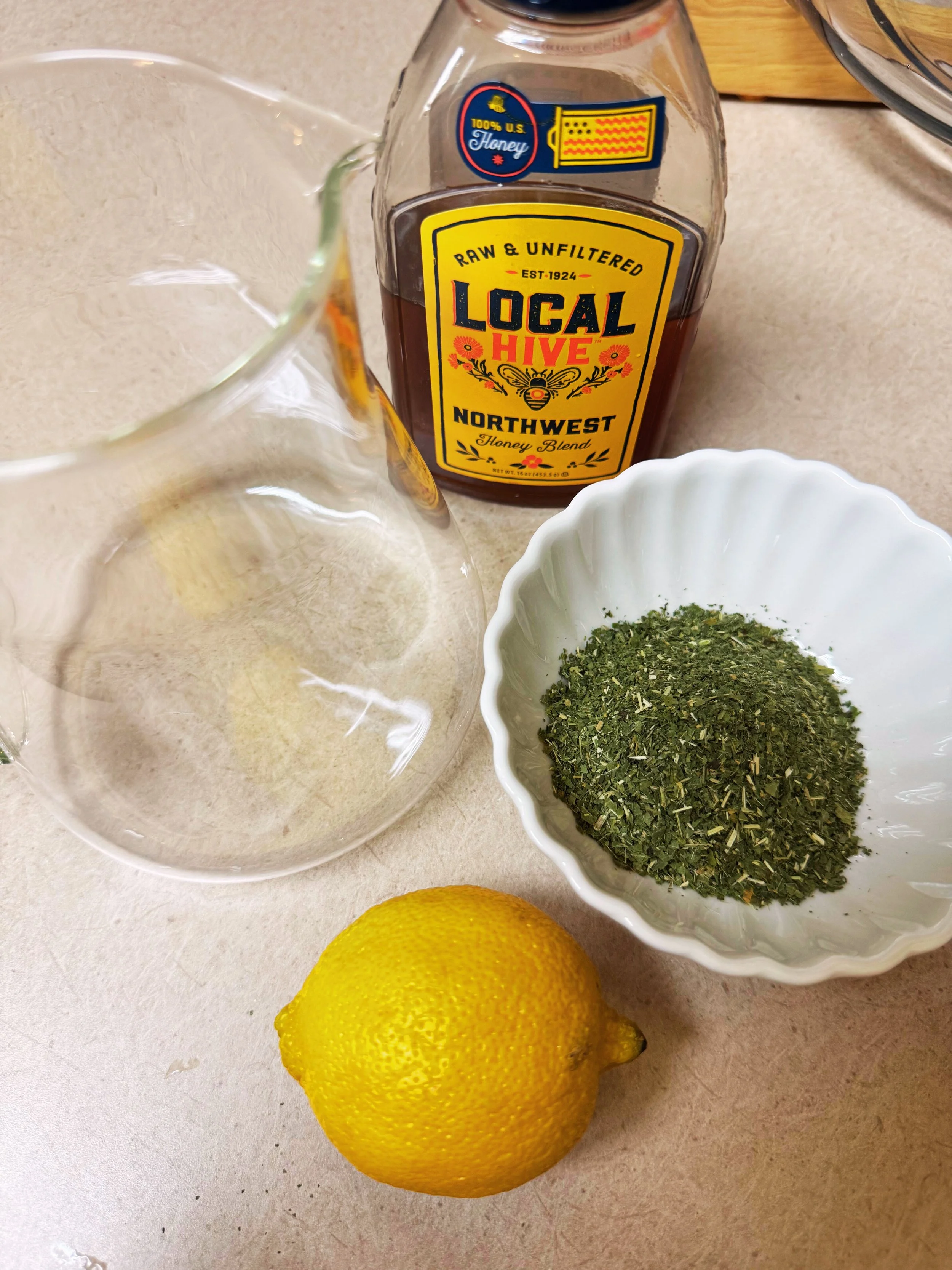 Ingredients for an Allergy Palmer: nettle, lemon, and local raw honey. Nettles are green powder in a white bowl, honney in bottle labelled northwest, and empty glass jar and a whole lemon