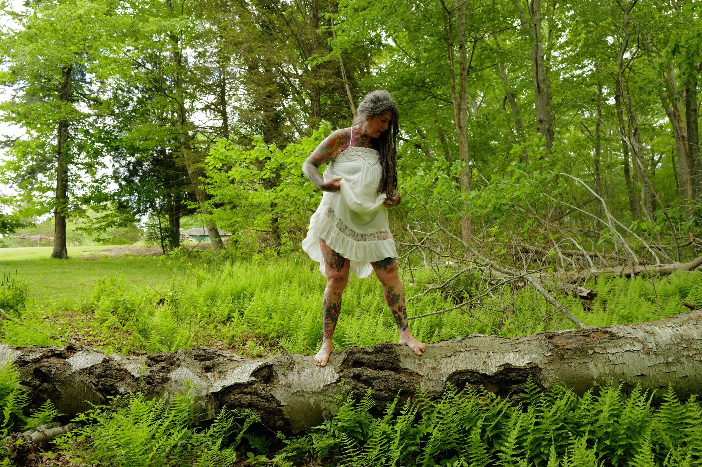 A woman with tattoos on her arms, legs, and chest, wearing a white dress, standing on a fallen tree over a creek in a lush green forest.