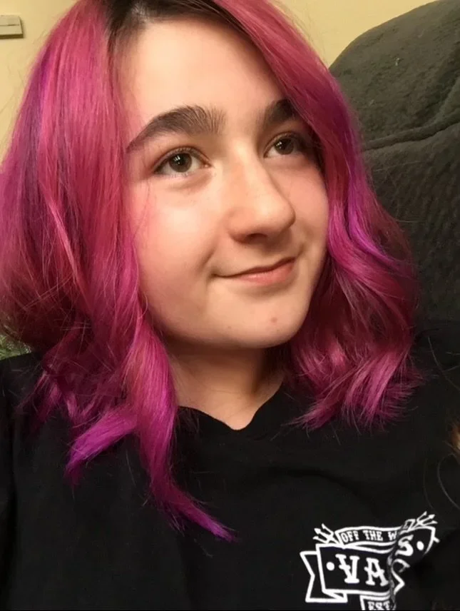 Young woman with pink and purple wavy hair smiling, wearing a black shirt with white text.