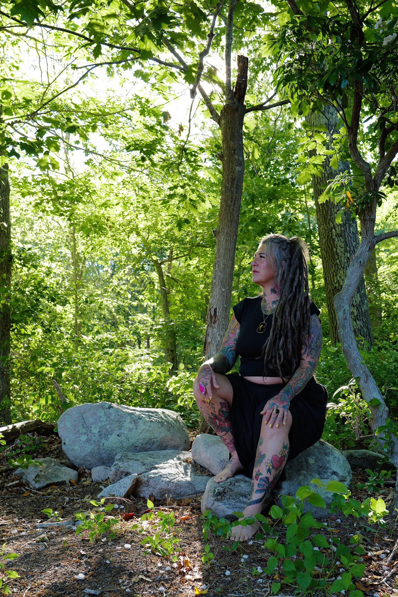 A woman with long dreadlocks and colorful tattoos is sitting on rocks in a lush green forest. She is wearing a black dress and appears to be looking to her left with a peaceful expression. Bright sunlight filters through the trees, illuminating her and the surrounding foliage.