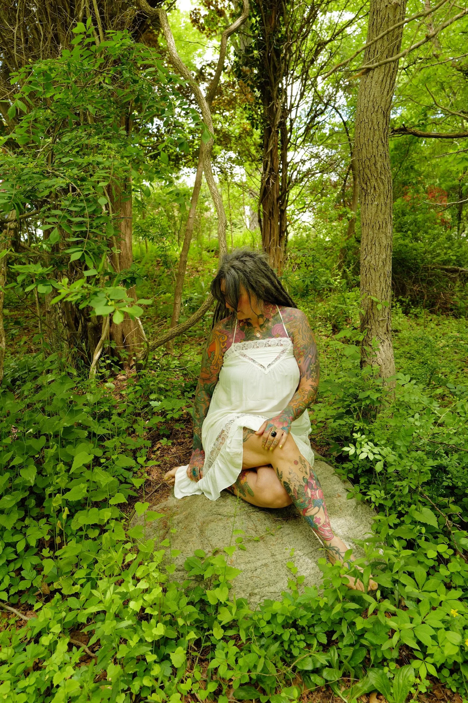 A woman with colorful tattoos and dreadlocks sitting on a stone in a lush green forest, wearing a white dress and holding her leg.