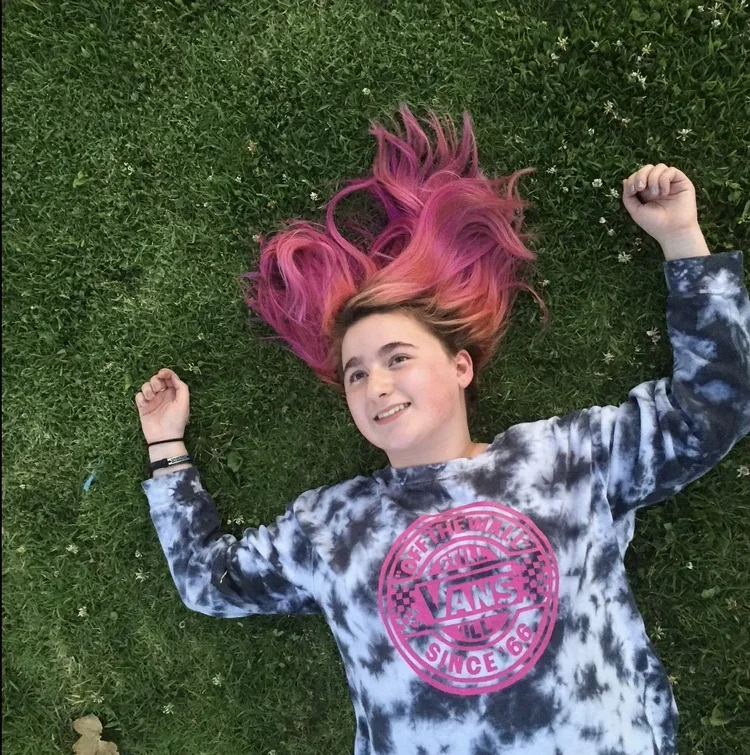 A girl with pink hair lying on green grass, smiling and looking up, wearing a black and white tie-dye Vans sweatshirt.