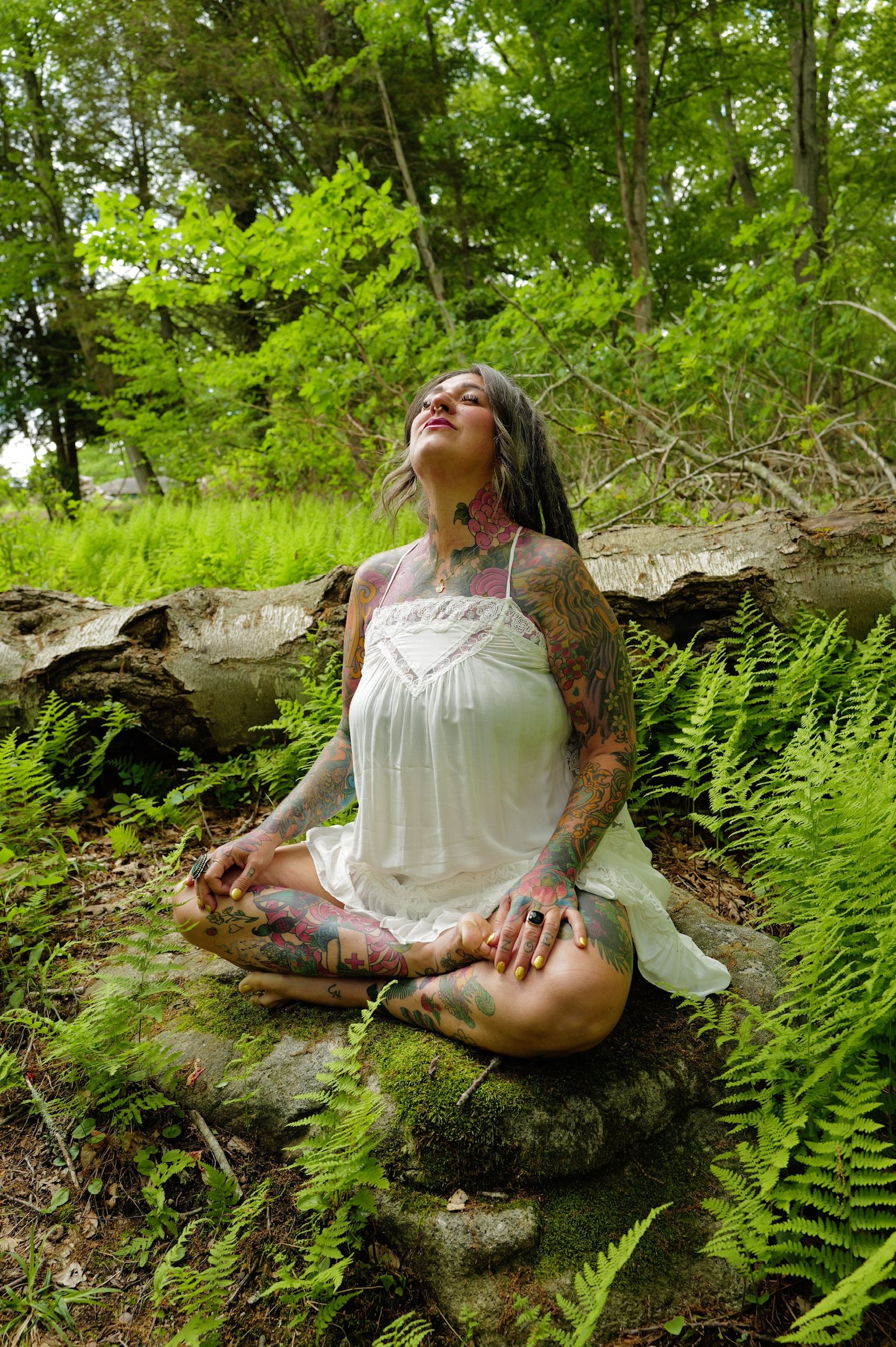 A woman with tattoos practicing meditation outdoors in a forest, sitting cross-legged on a moss-covered log, with a serene expression and surrounded by green foliage.