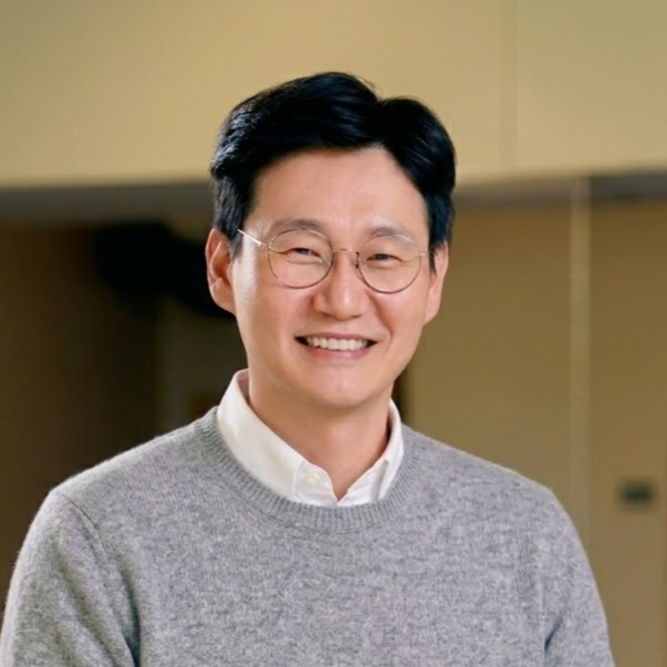 Close-up of a smiling man with dark hair, glasses, wearing a black blazer and a striped shirt, in a bright indoor space with windows in the background.