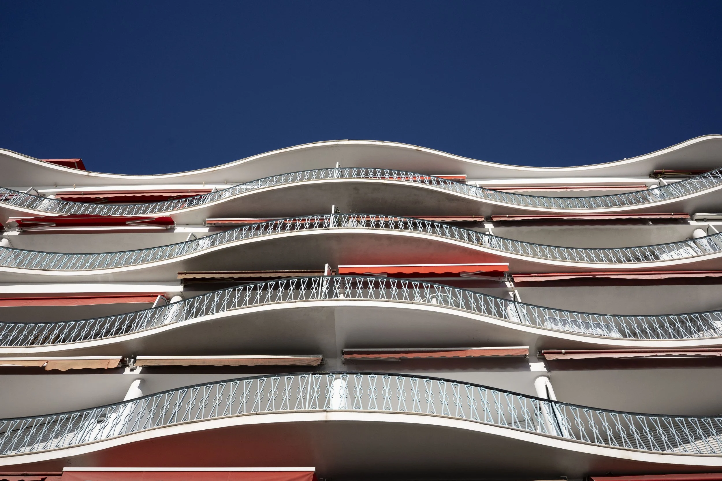 Mehrstöckiges Gebäude mit geschwungenen Balkonen und roten Markisen gegen einen klaren blauen Himmel.