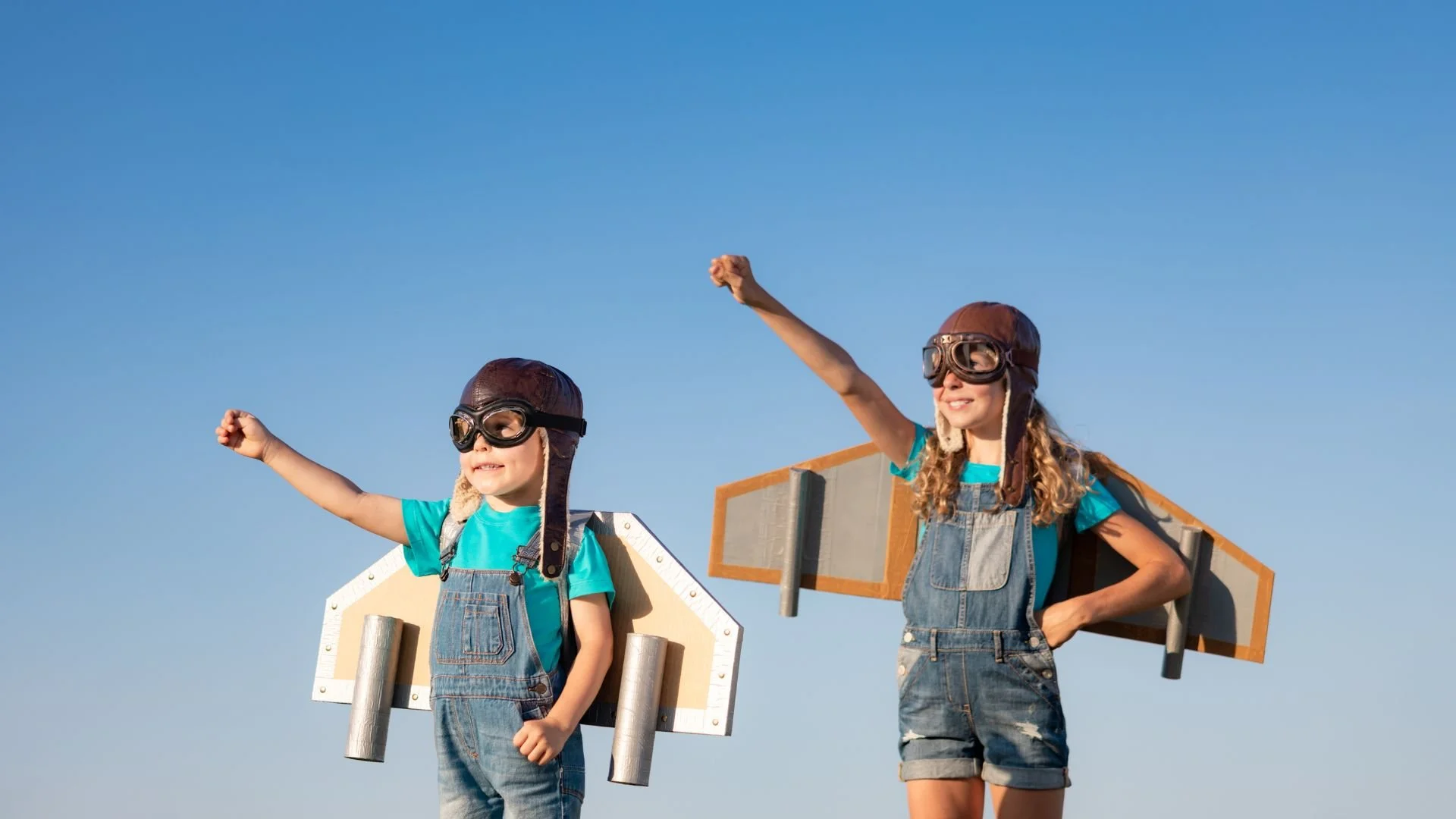 Two children wearing aviator hats and goggles, with cardboard jet pack wings, pretending to fly against a clear blue sky.