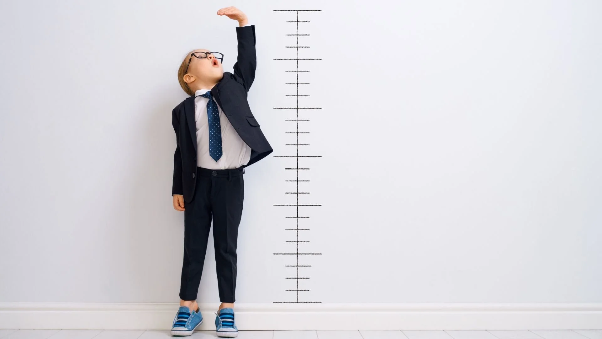 Young boy in a suit reaching up beside a height measurement chart on a wall.