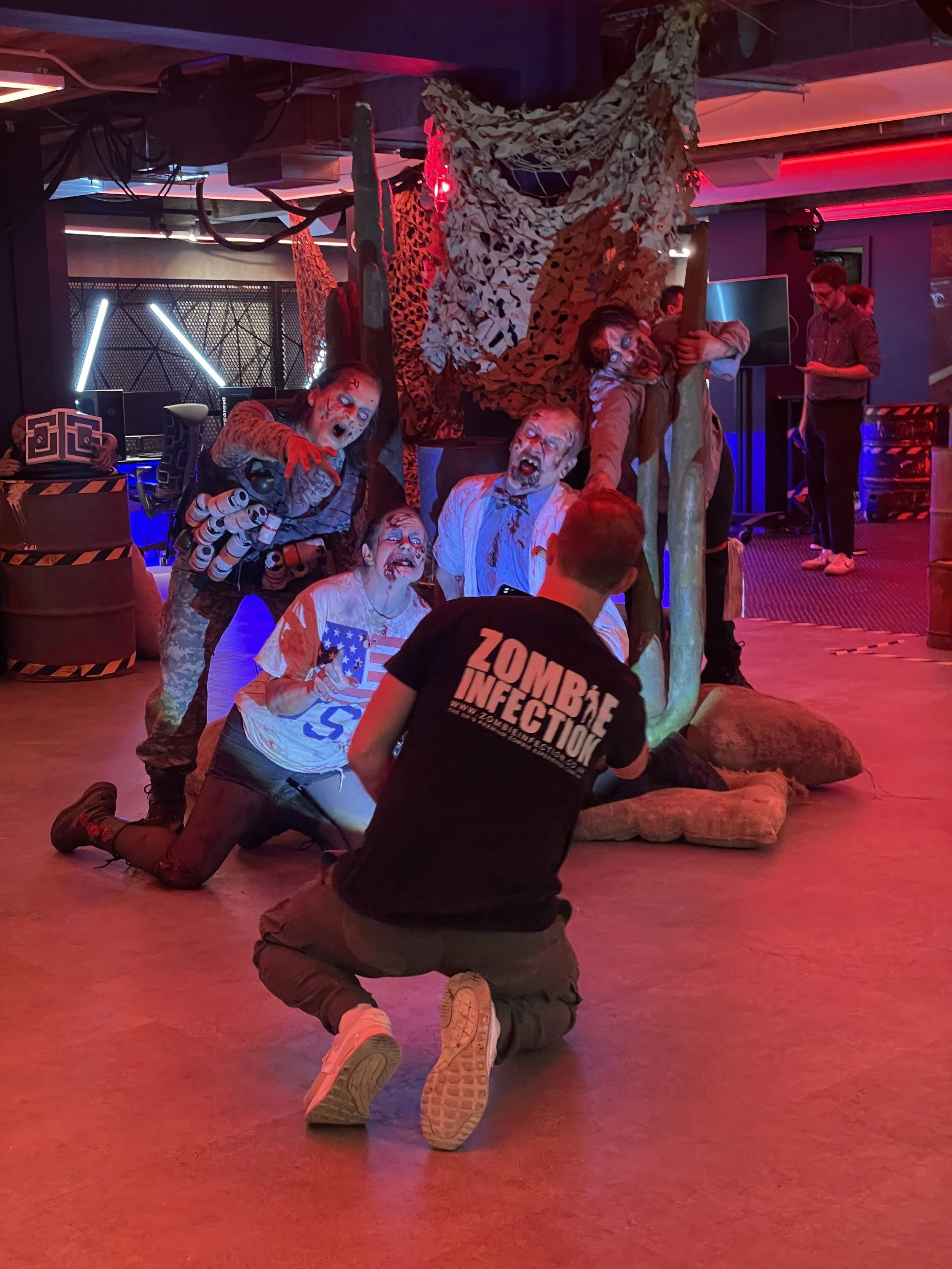 A group of people dressed as zombies with makeup and costumes are posing for a photo in a dark, decorated setting. One person is kneeling in front, wearing a "Zombie Infection" shirt, while others are around a tree and camouflage netting, all with zo