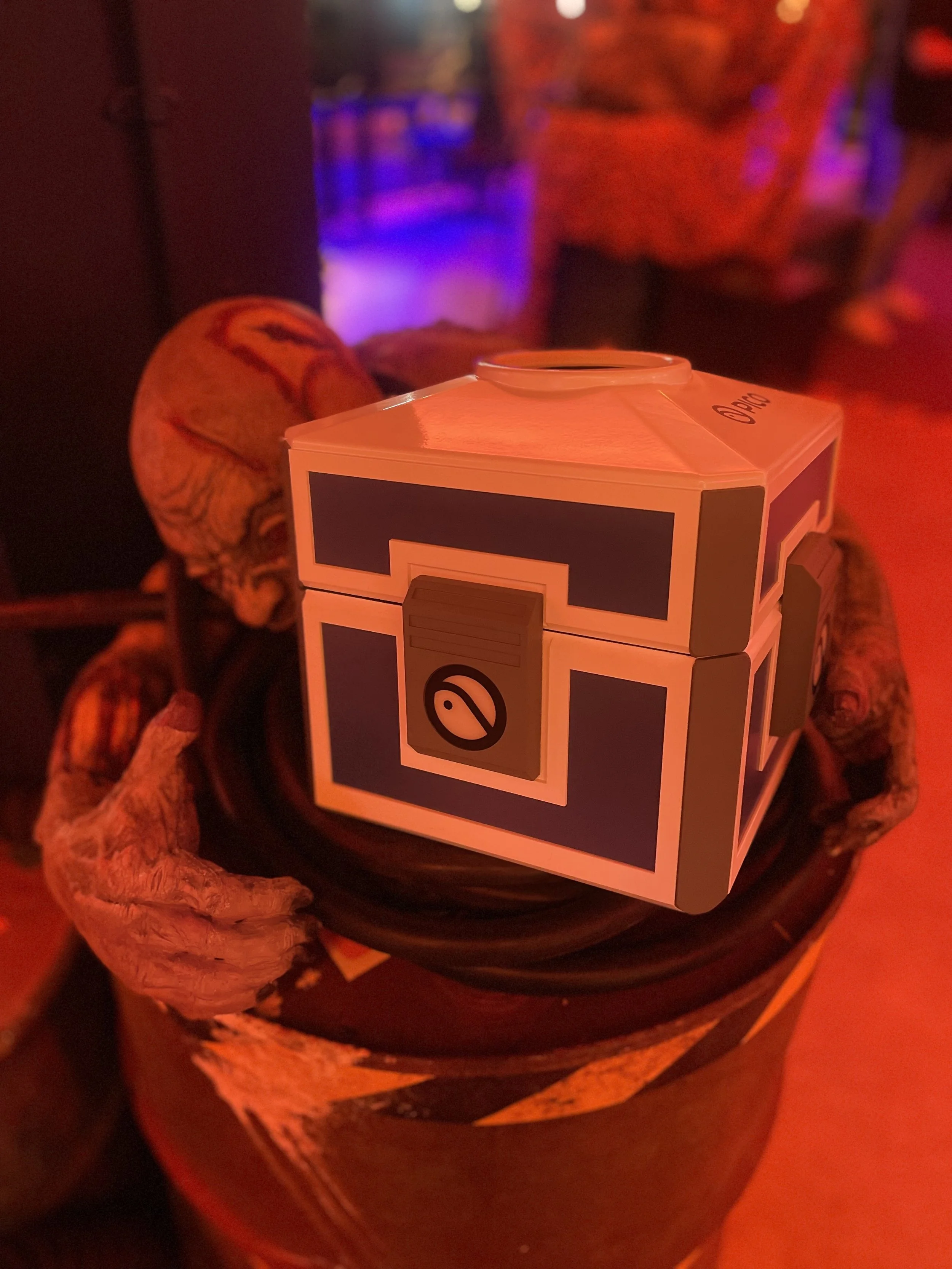 A cardboard box designed to resemble a treasure chest, sitting on top of a dustbin with a clawed animal's paw holding the dustbin. The background has a reddish hue and some purple and blue lighting, indicating a club or bar setting.
