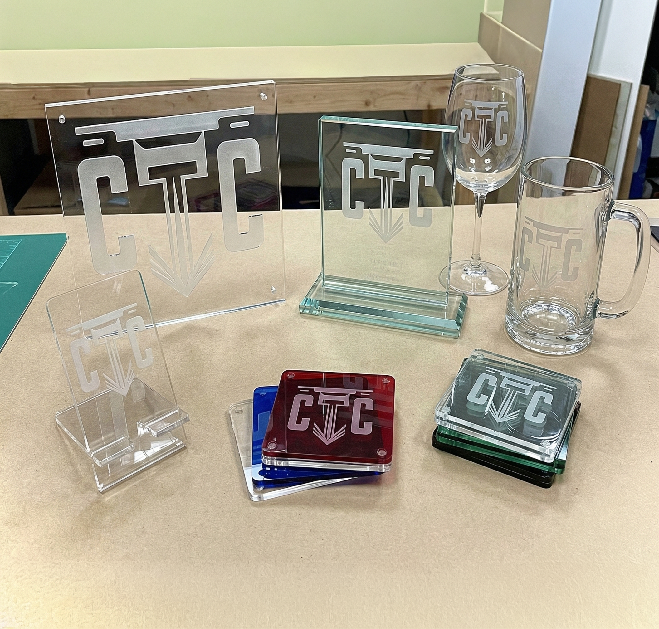 Various glass and acrylic display stands, coasters, and glassware with a logo that has the letters 'CTC'  design etched on them, arranged on a beige table.