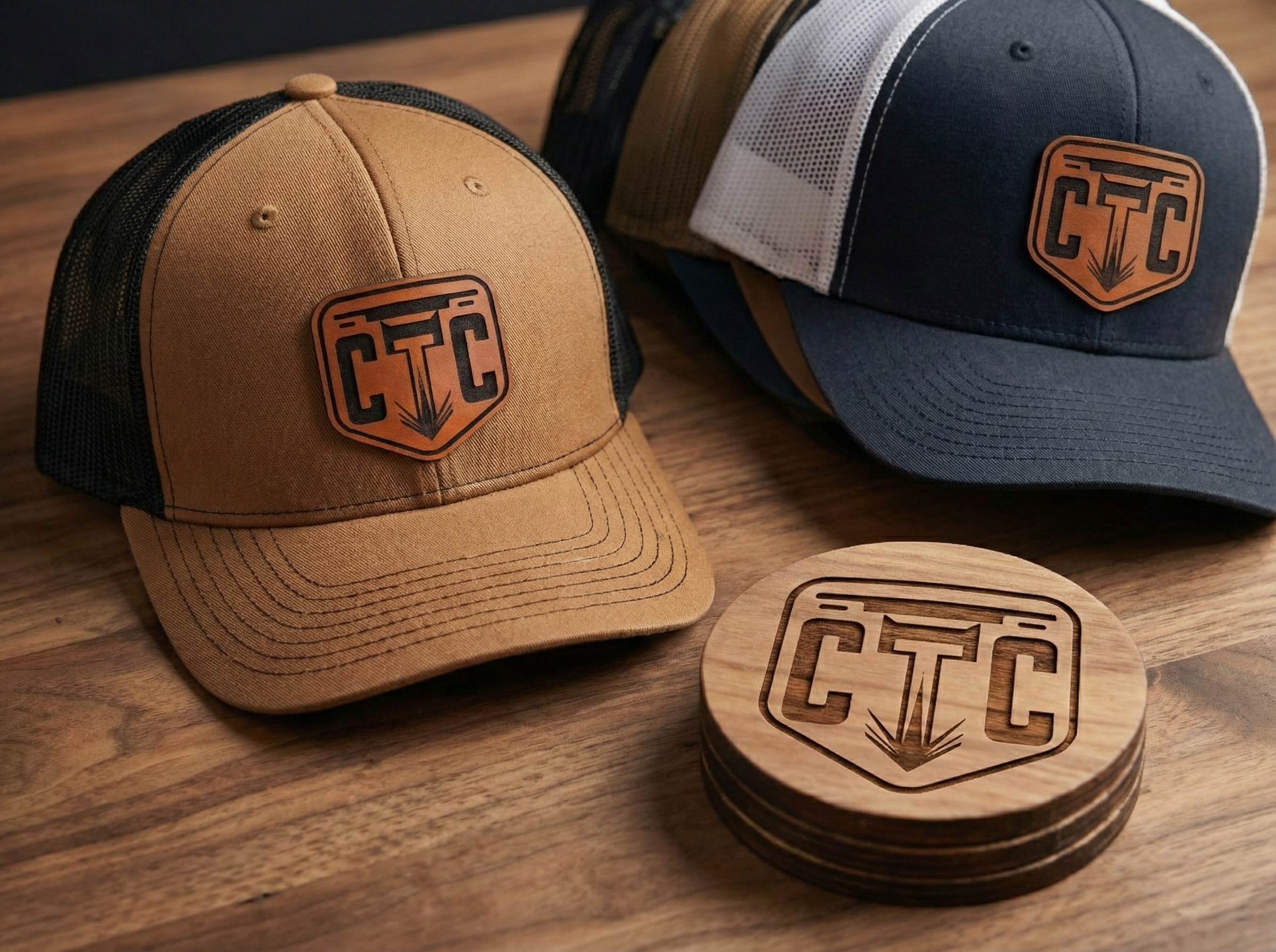 Two baseball caps, one tan and black, one blue and gray, with a company branding logo patch on the front. There are also coasters with the same company logo on a wooden surface.