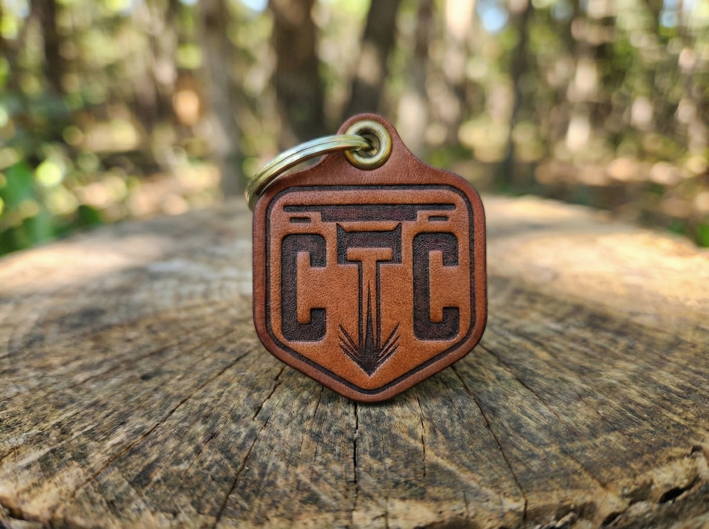 Leather keychain with engraved geometric design, placed on a weathered wooden surface outdoors with blurred trees in the background.