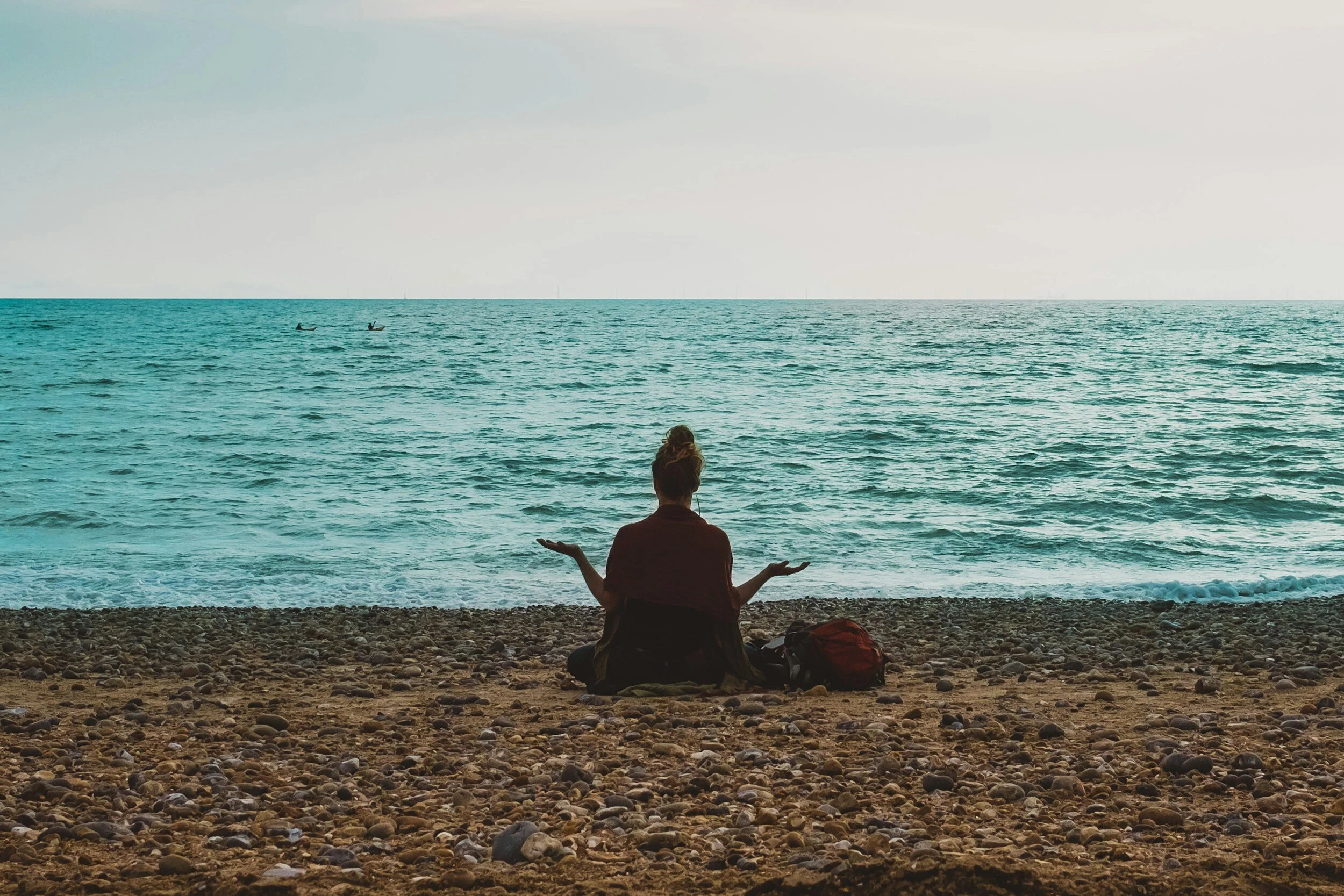 A person sitting cross-legged on a pebble beach, facing the ocean with arms raised and hands open in a meditative pose, with two backpacks beside them and boats in the distant water.
