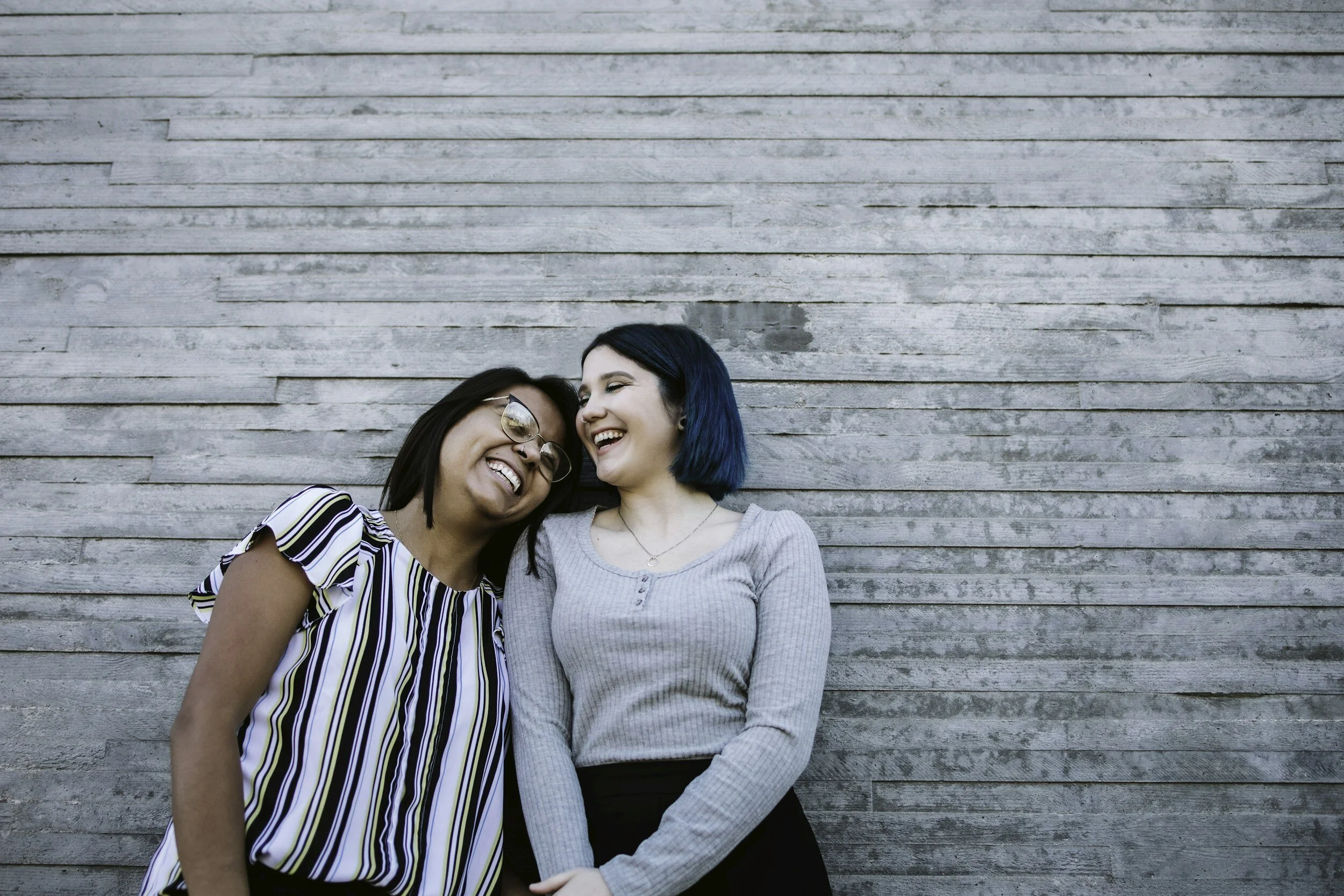 Two women laughing and smiling leaning against a weathered wooden wall.