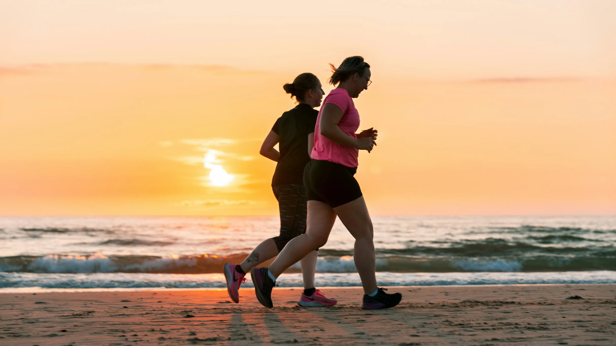 Two women jogging on the beach at sunset with waves in the background.