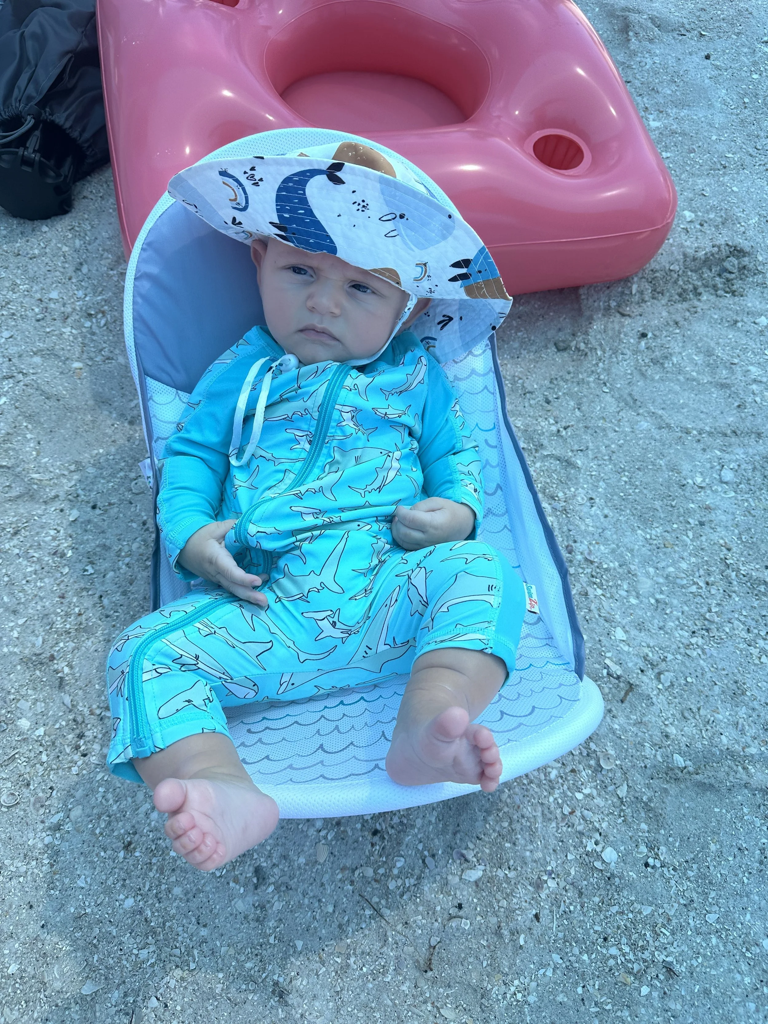 Baby in a green zip-up shark rash guard on a beach towel