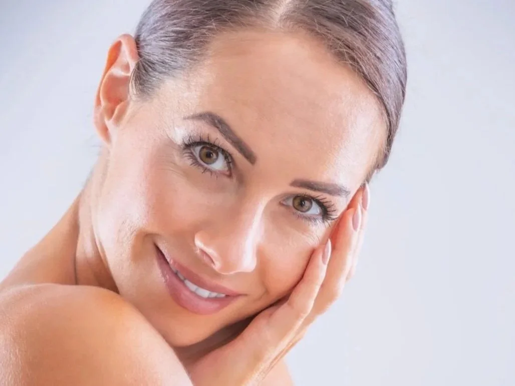 Close-up of a smiling woman with clear skin and brown hair, resting her face in her hand.